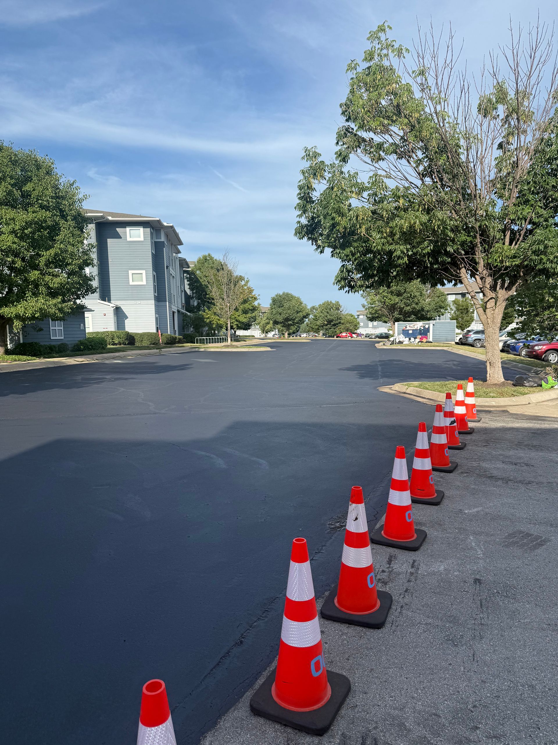 A row of orange traffic cones separates newly paved black asphalt from a grey, textured parking lot beside apartment units.