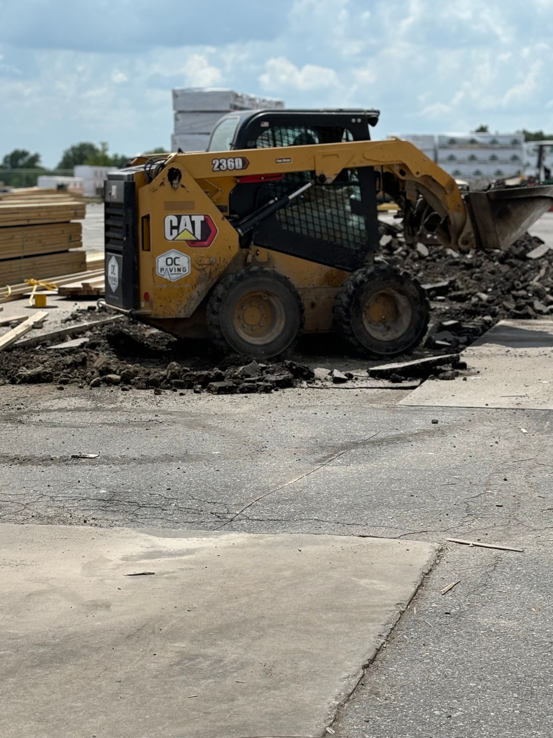 A yellow Caterpillar skid steer loader sits on a dirt patch at a construction site with stacks of lumber nearby.