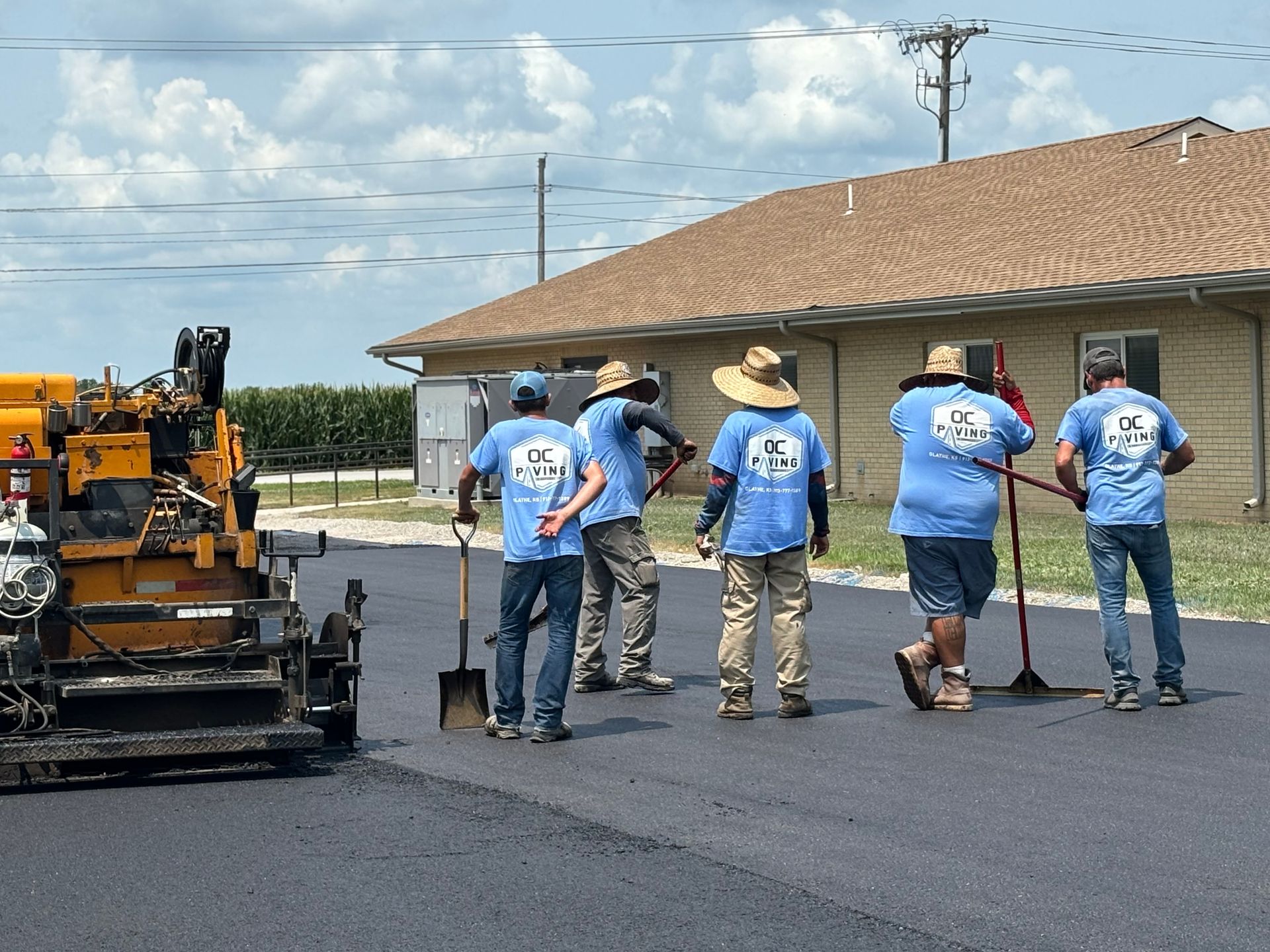 Five workers in blue shirts and hats use paving tools to spread asphalt next to a construction vehicle and a building.
