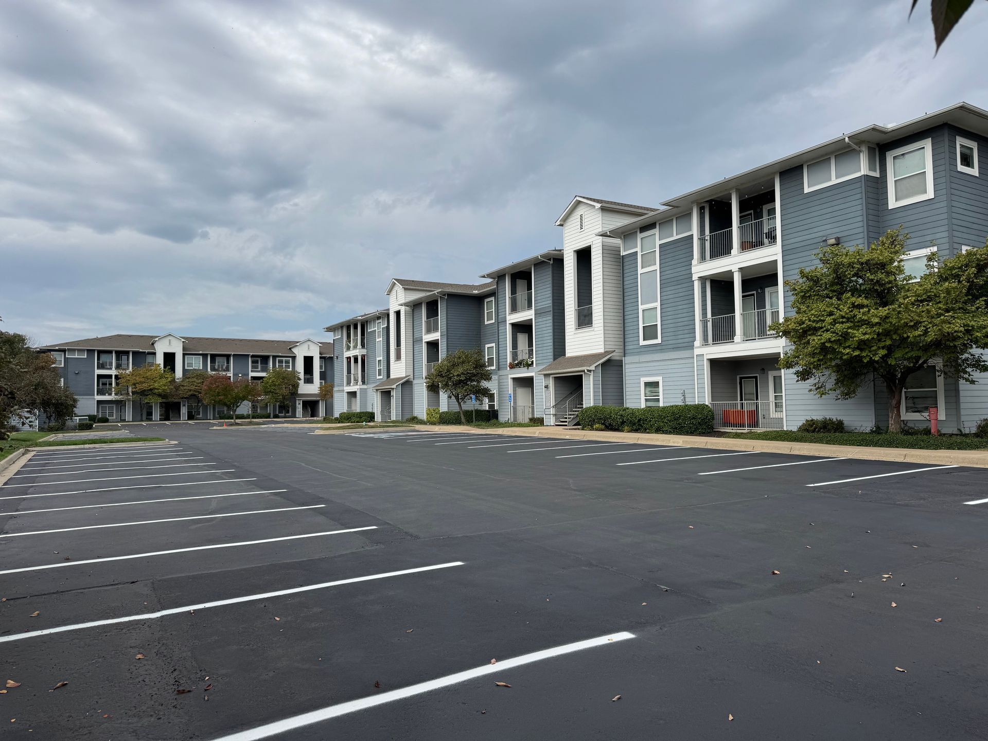 A multi-story apartment building with blue-gray siding, balconies, and a large, empty asphalt parking lot under a cloudy sky.