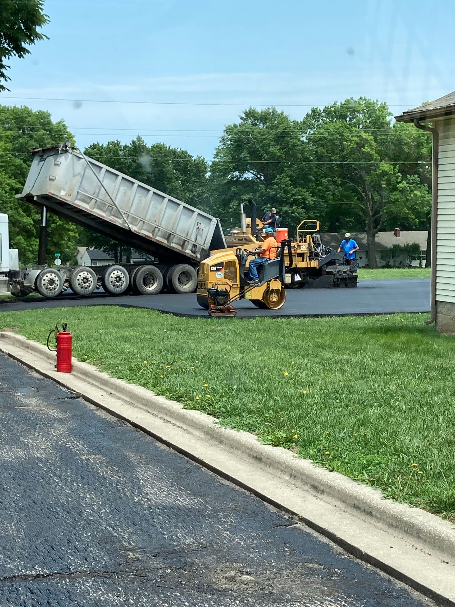 A dump truck is unloading asphalt into a paving machine on a construction site, with workers nearby on a sunny day.