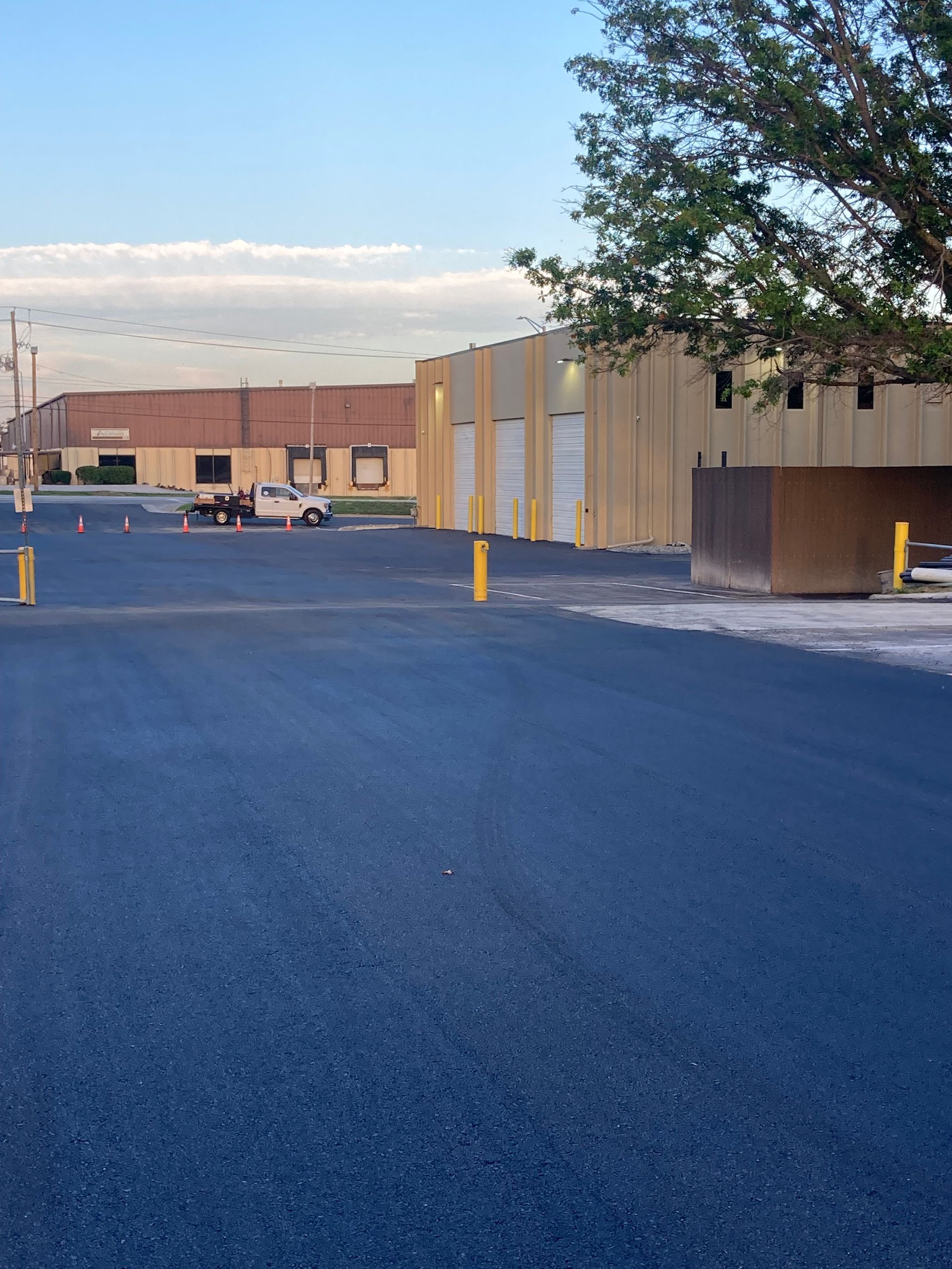 A wide view of a paved parking lot facing a tan industrial building with bay doors and a truck parked in the distance.