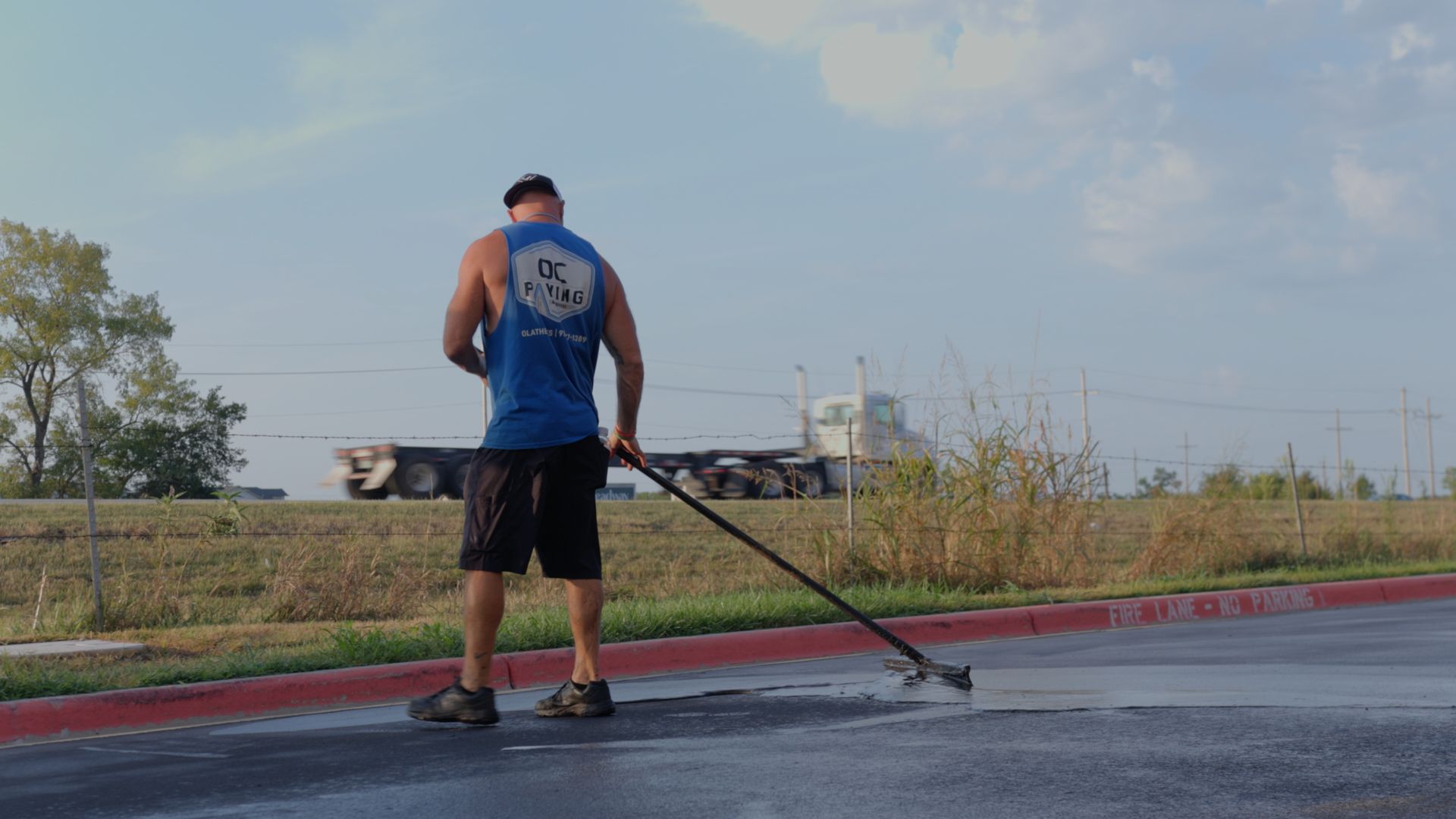 A person in a blue tank top and black shorts uses a long-handled tool to spread liquid sealant on a parking lot surface.