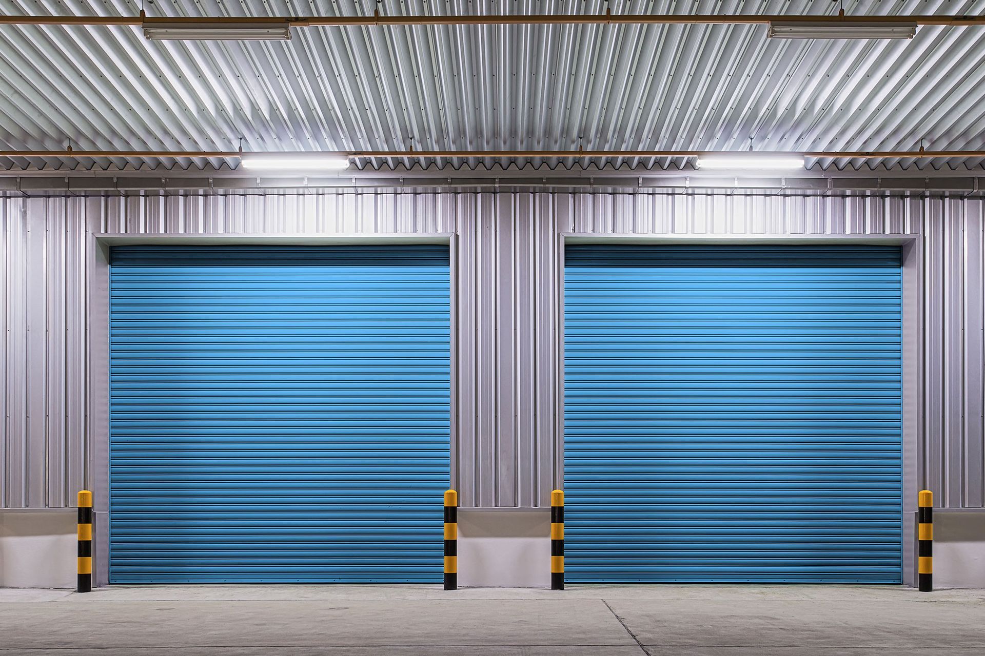 Two closed blue roll-up doors on a metal building with overhead lighting and bollards.