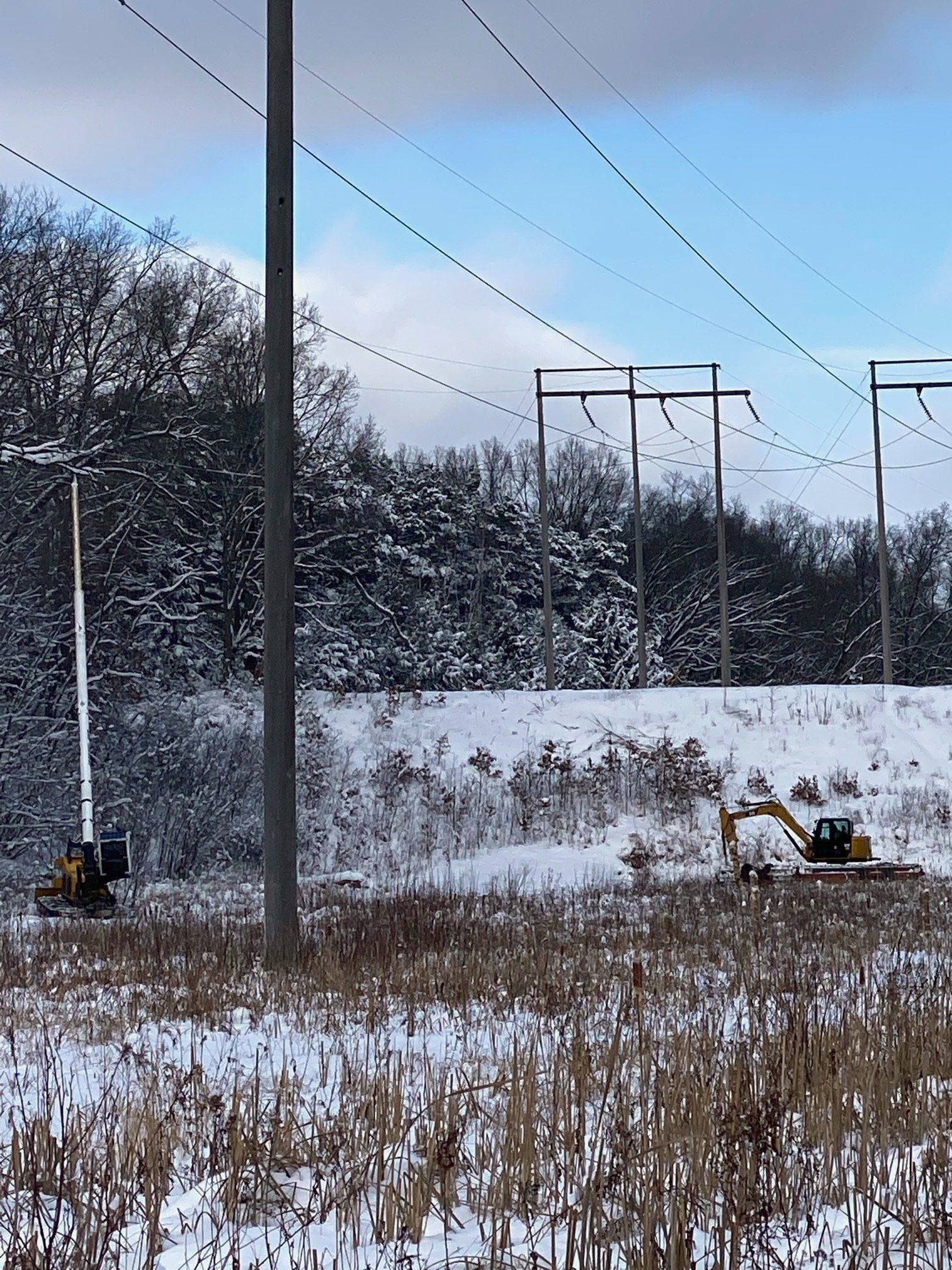 A yellow excavator is driving through a snowy field next to power lines.