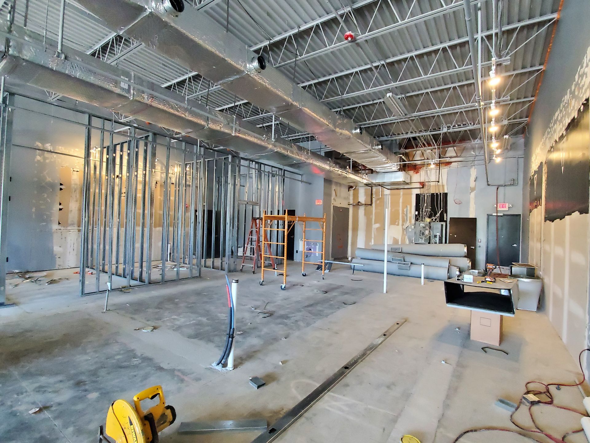 Interior of a building under construction, metal studs define walls, exposed ceiling, and tools on the concrete floor.
