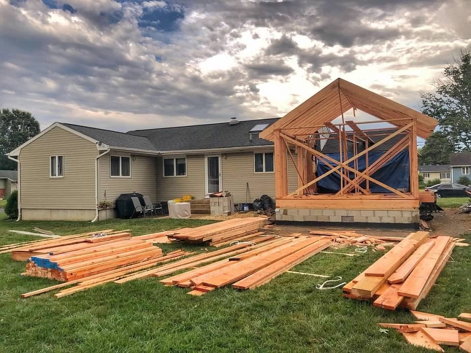 Backyard construction: framing an addition to a beige house. Lumber scattered on grass. Cloudy sky.