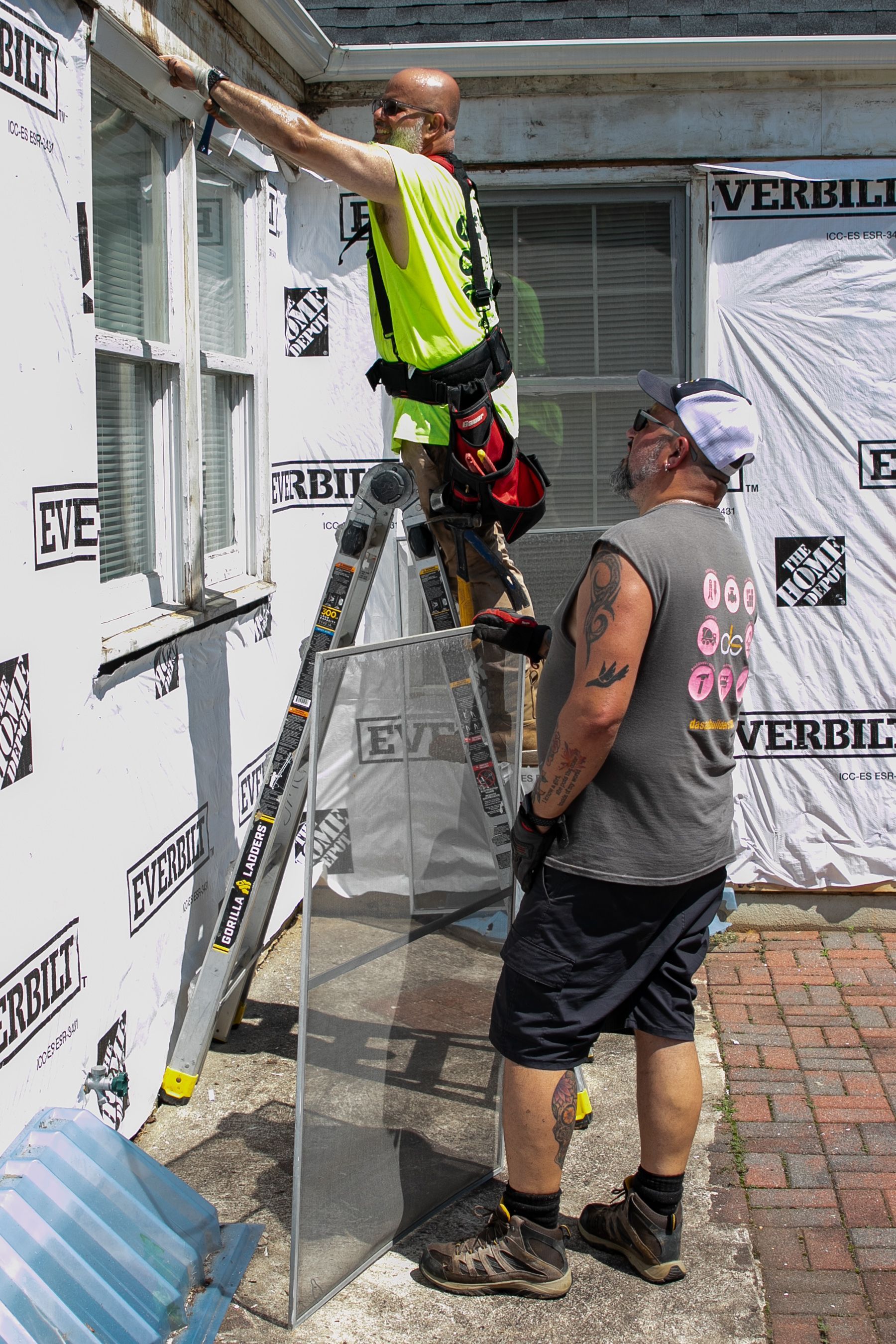 Two men work on a building exterior. One on ladder paints window trim. The other watches.