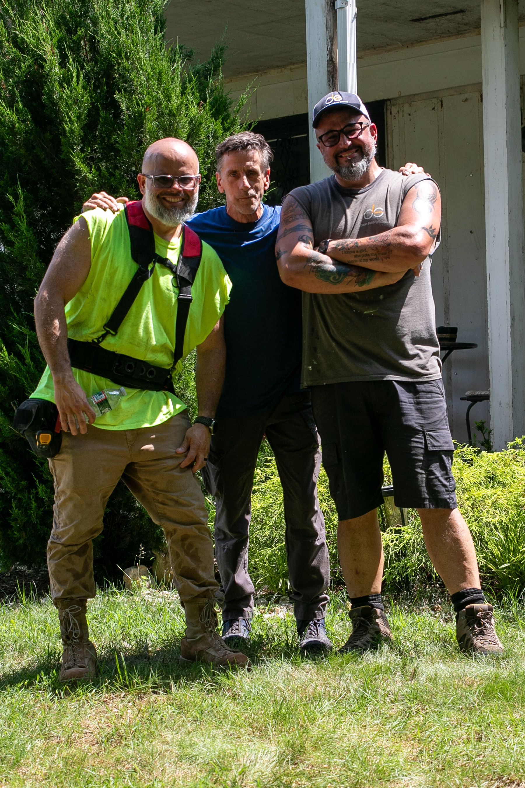 Three men in front of a house; one in neon vest, one with arms crossed, one looking at the camera.