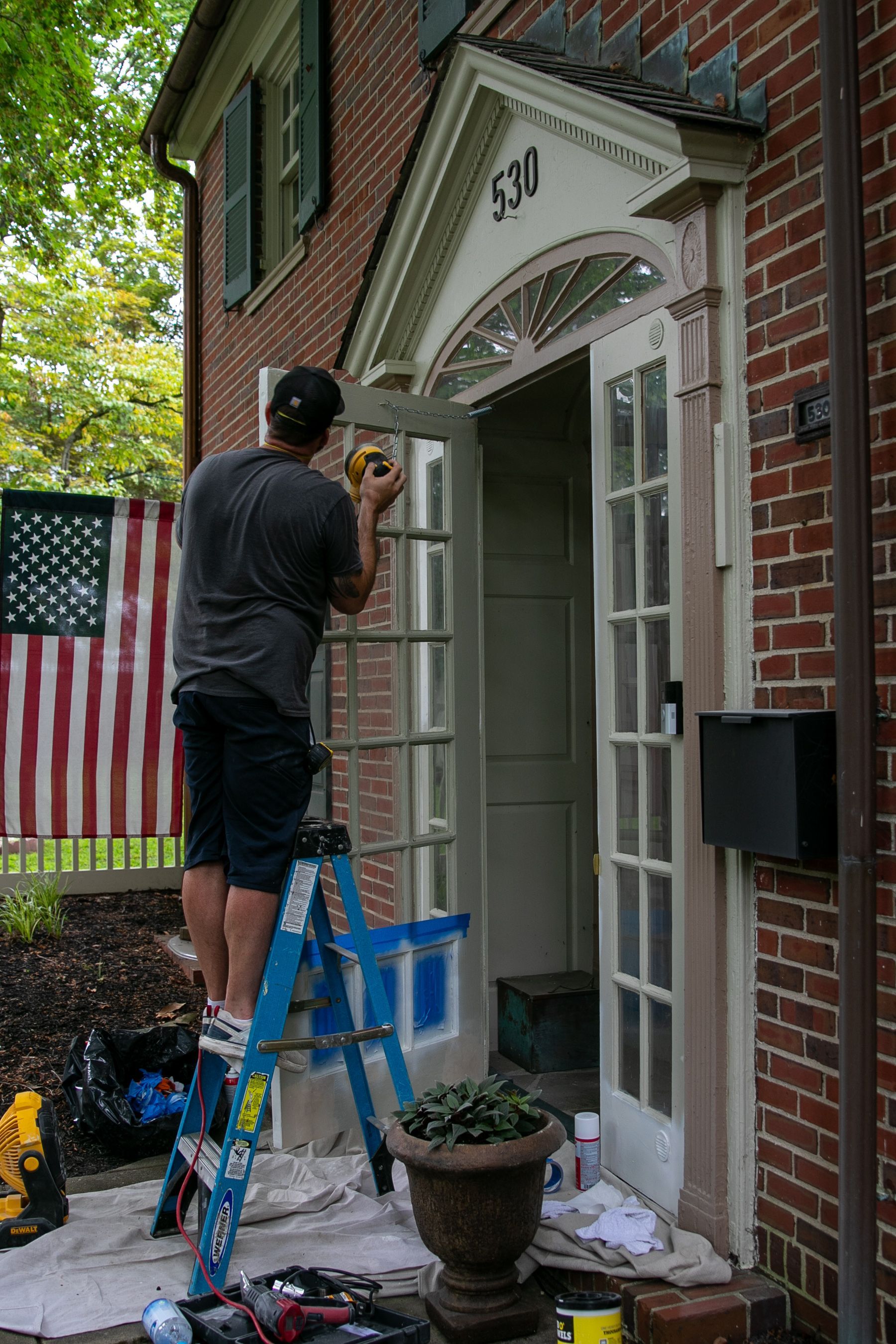Man on a ladder painting door frame of a brick house, with an American flag in the background.