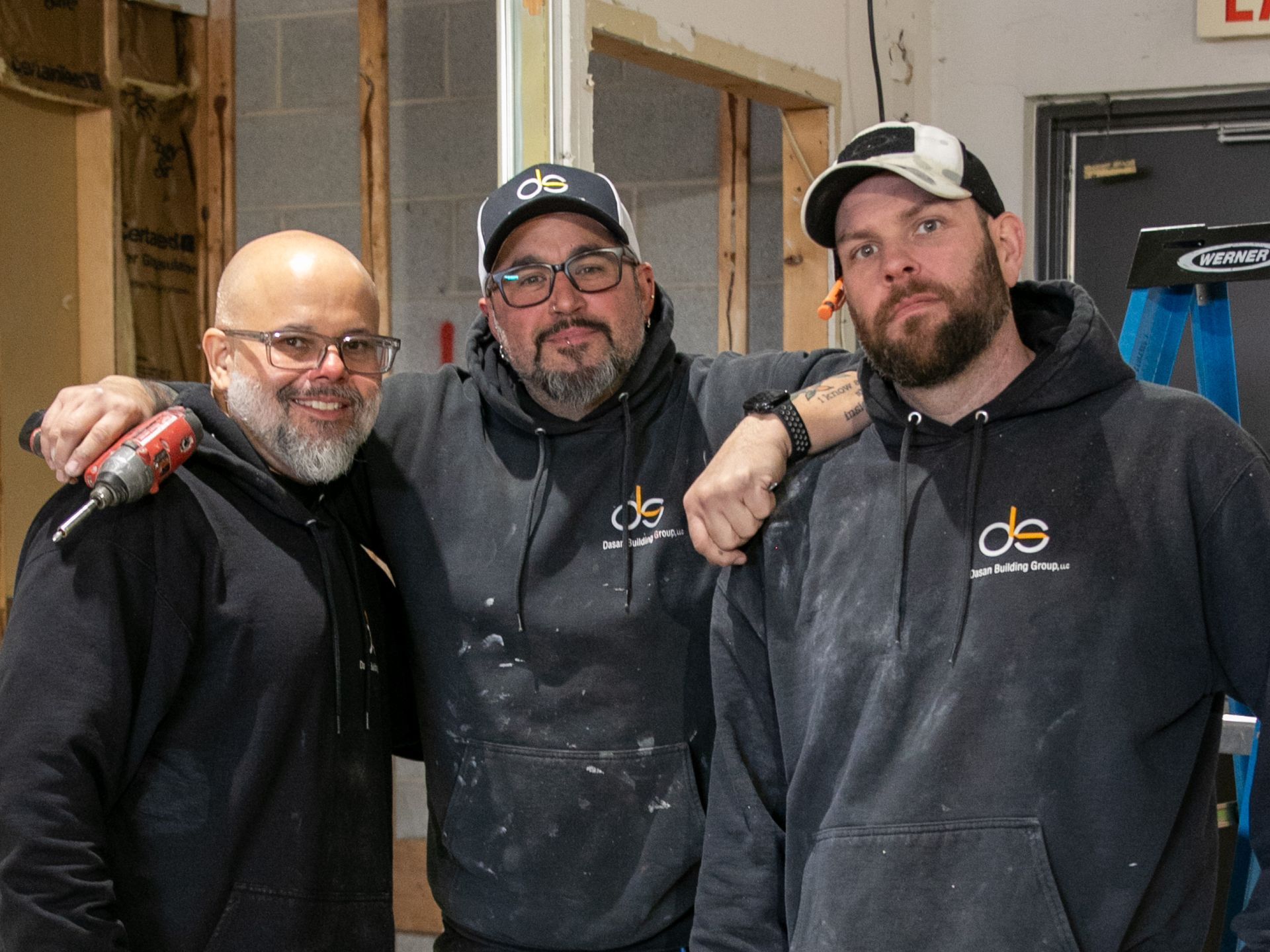 Three men in construction setting, posing with arms around each other, wearing matching hoodies.