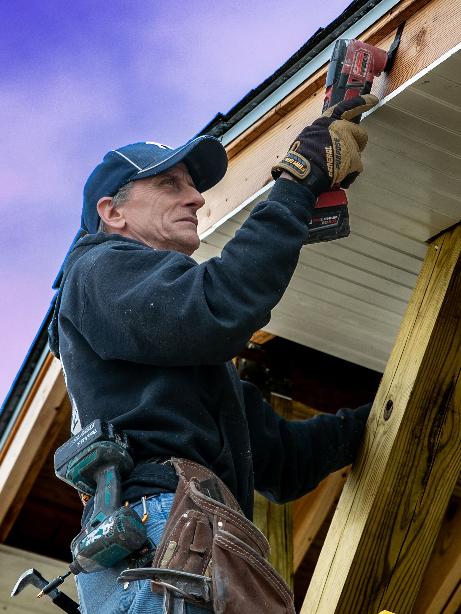 Man on ladder using a drill to install siding on a building, wearing a cap and work gloves.