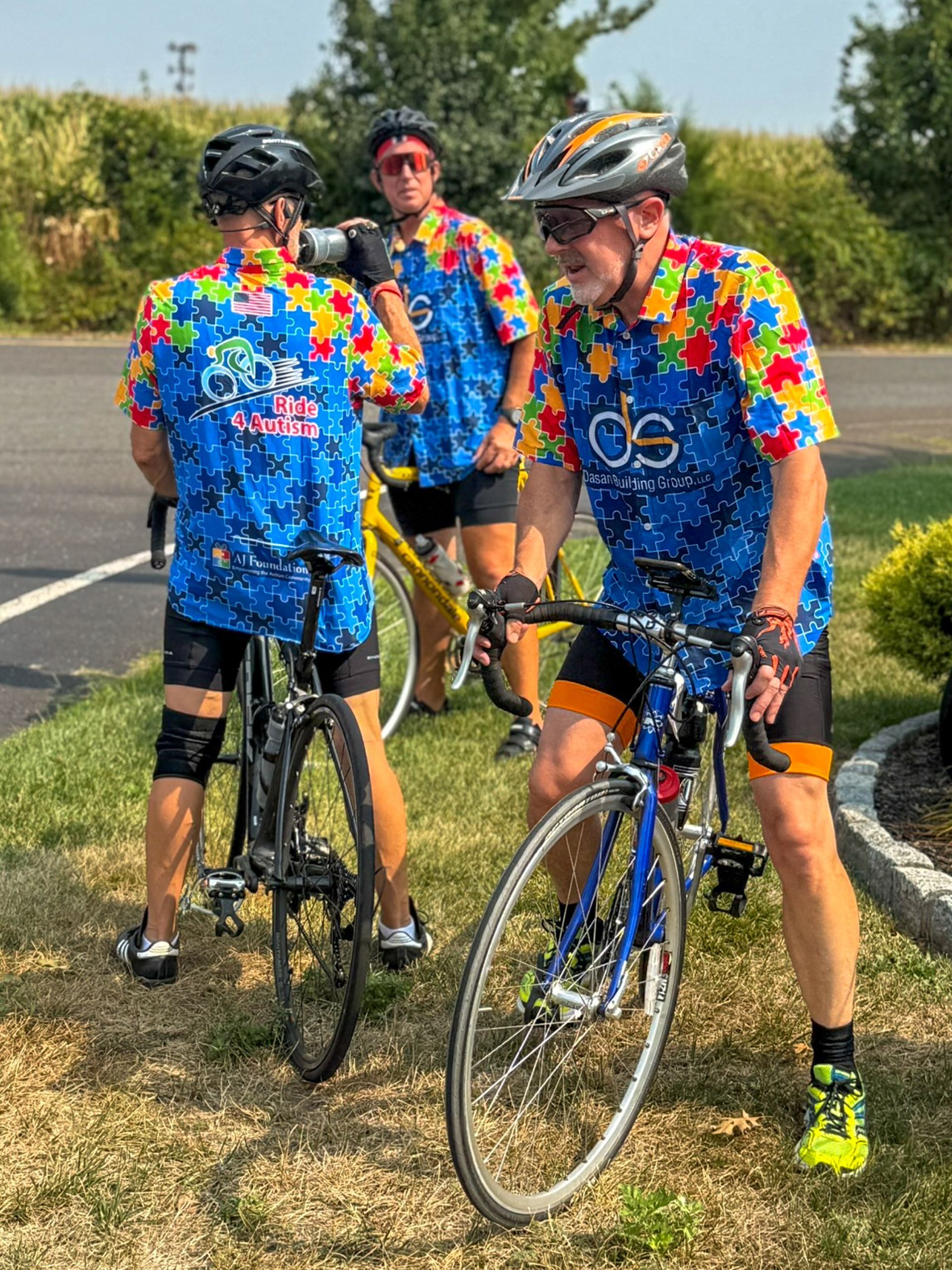 Three cyclists in blue puzzle-piece shirts with bikes, posing outdoors on a sunny day.