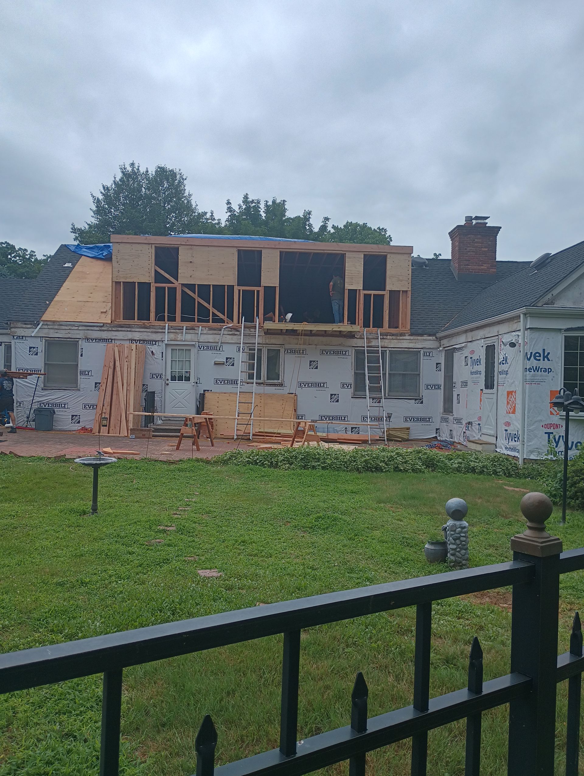 House under construction, with new wooden upper level framed above existing white siding. Overcast day.