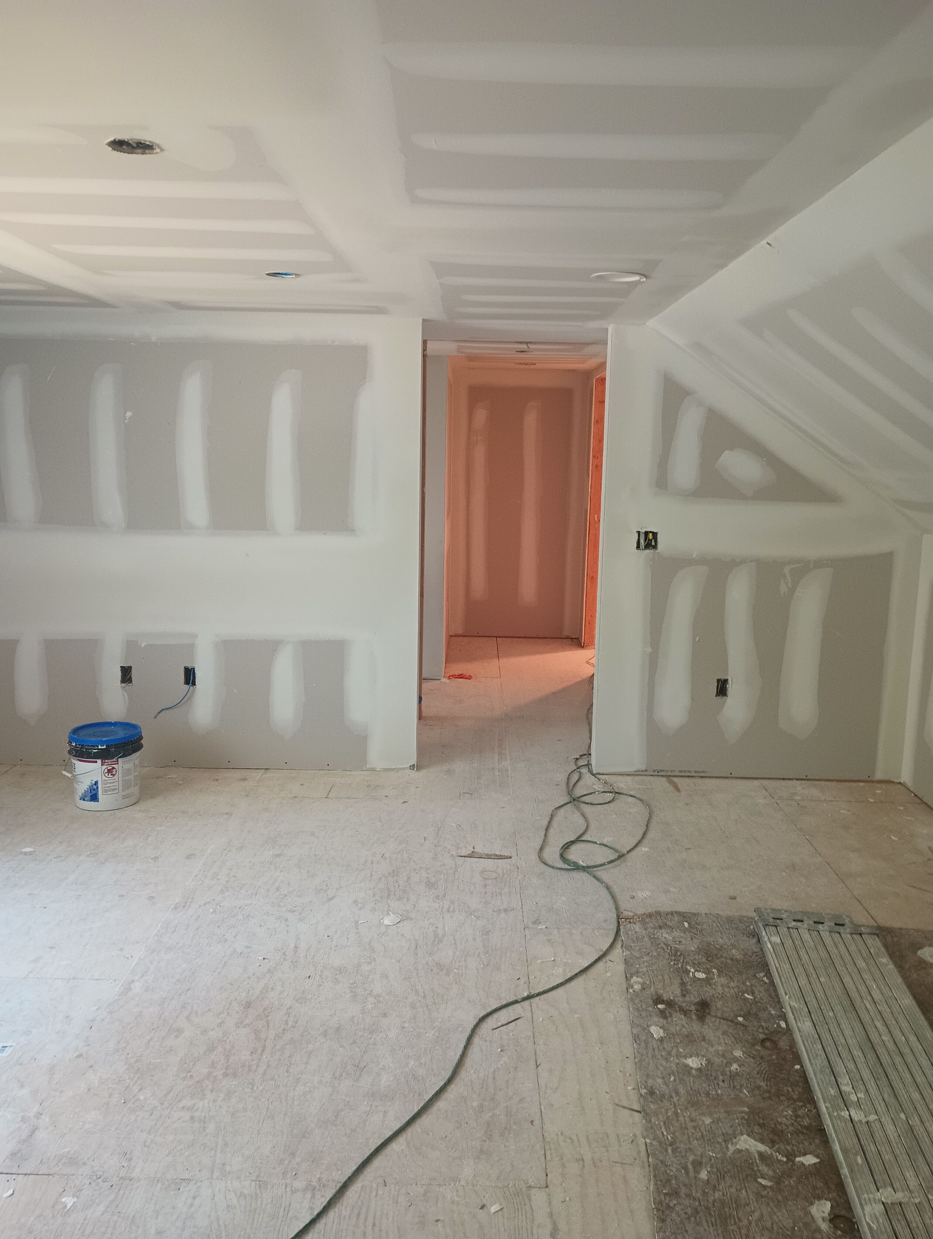 Interior view of a room under construction, with drywall on walls and ceiling; open doorway leads to another room.