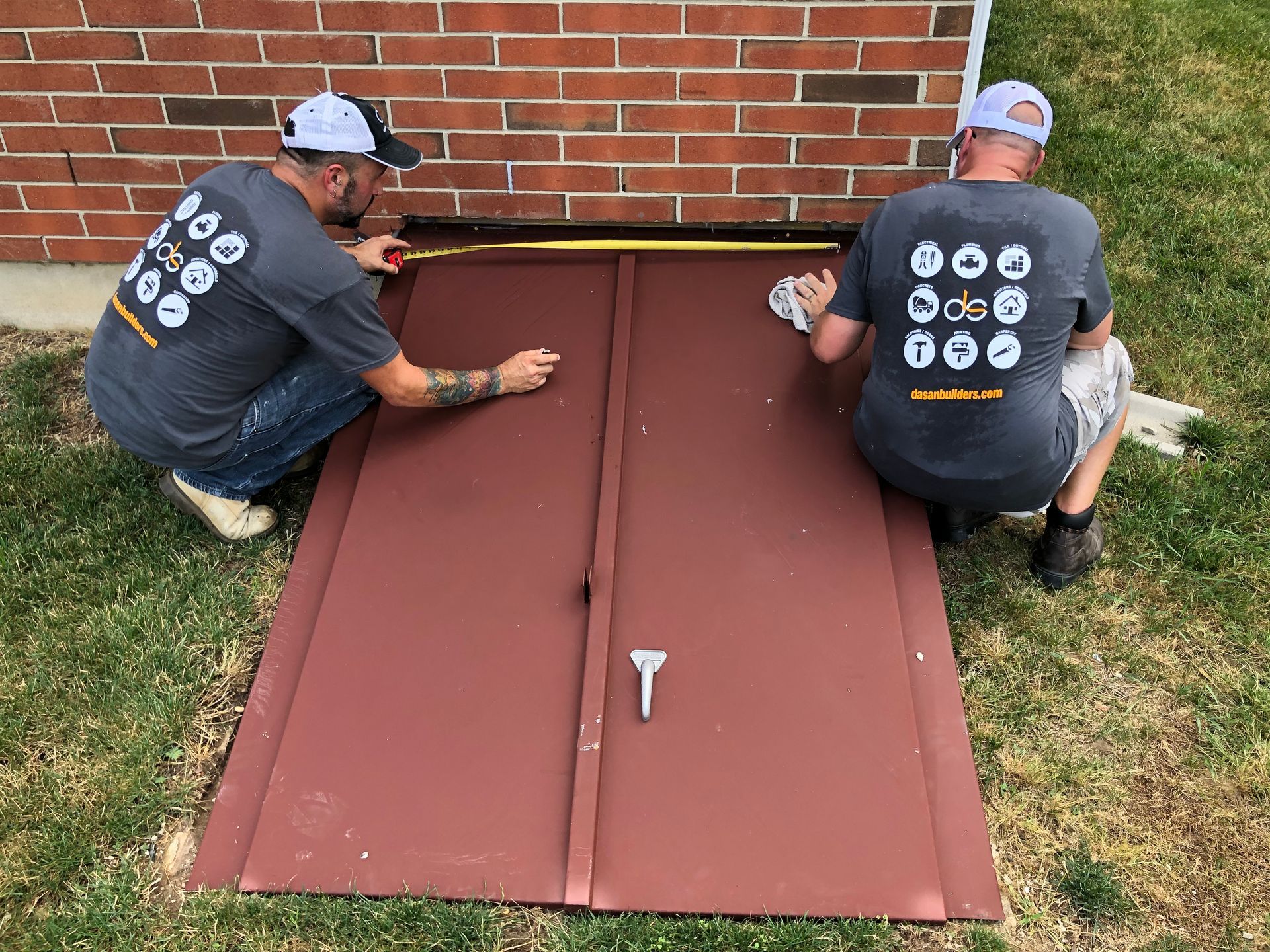 Two men in gray shirts measure and work on a maroon basement door against a brick wall, outside on grass.