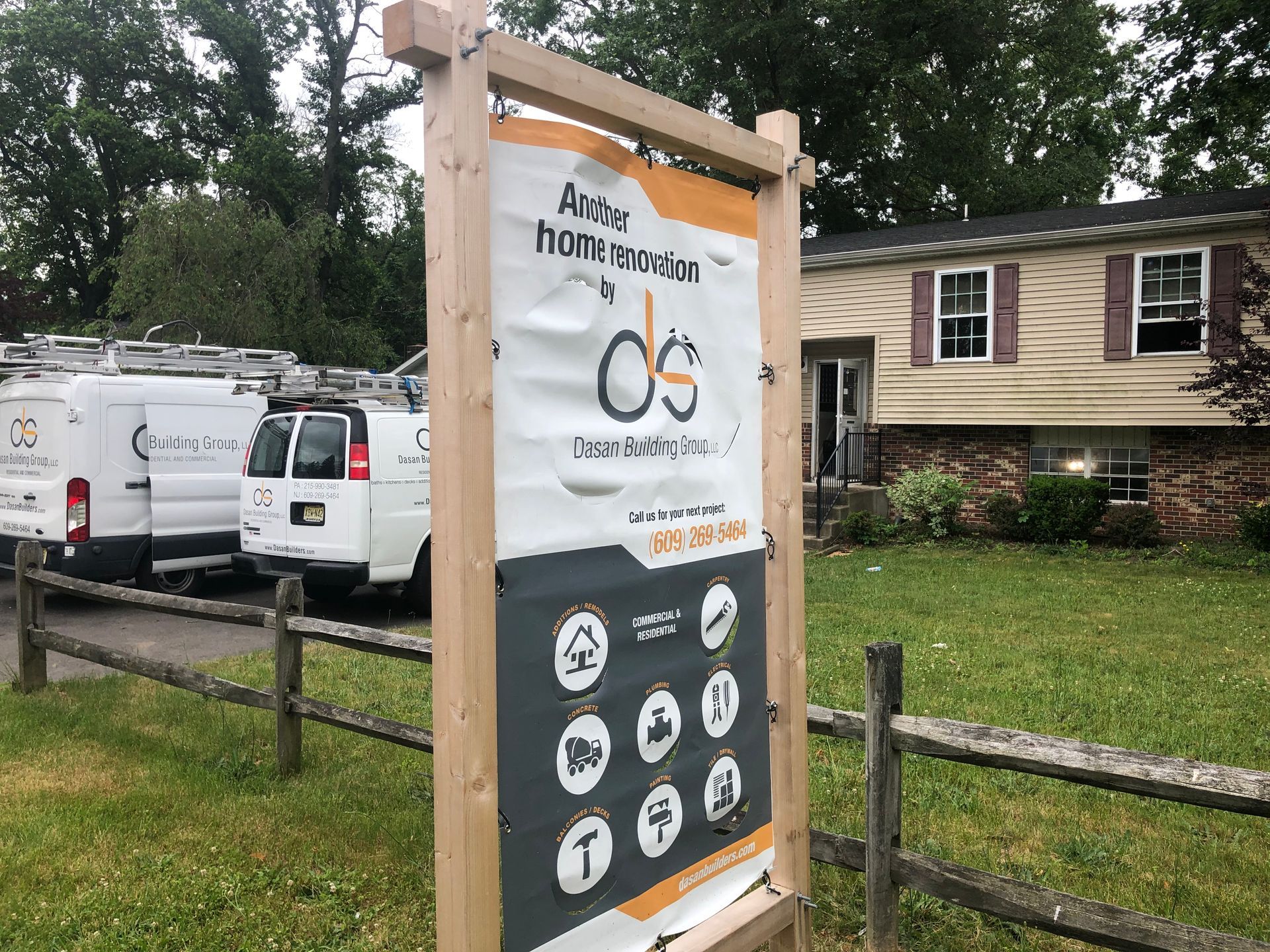 A home renovation sign stands in front of a split-level house with vans parked beside a wooden fence.