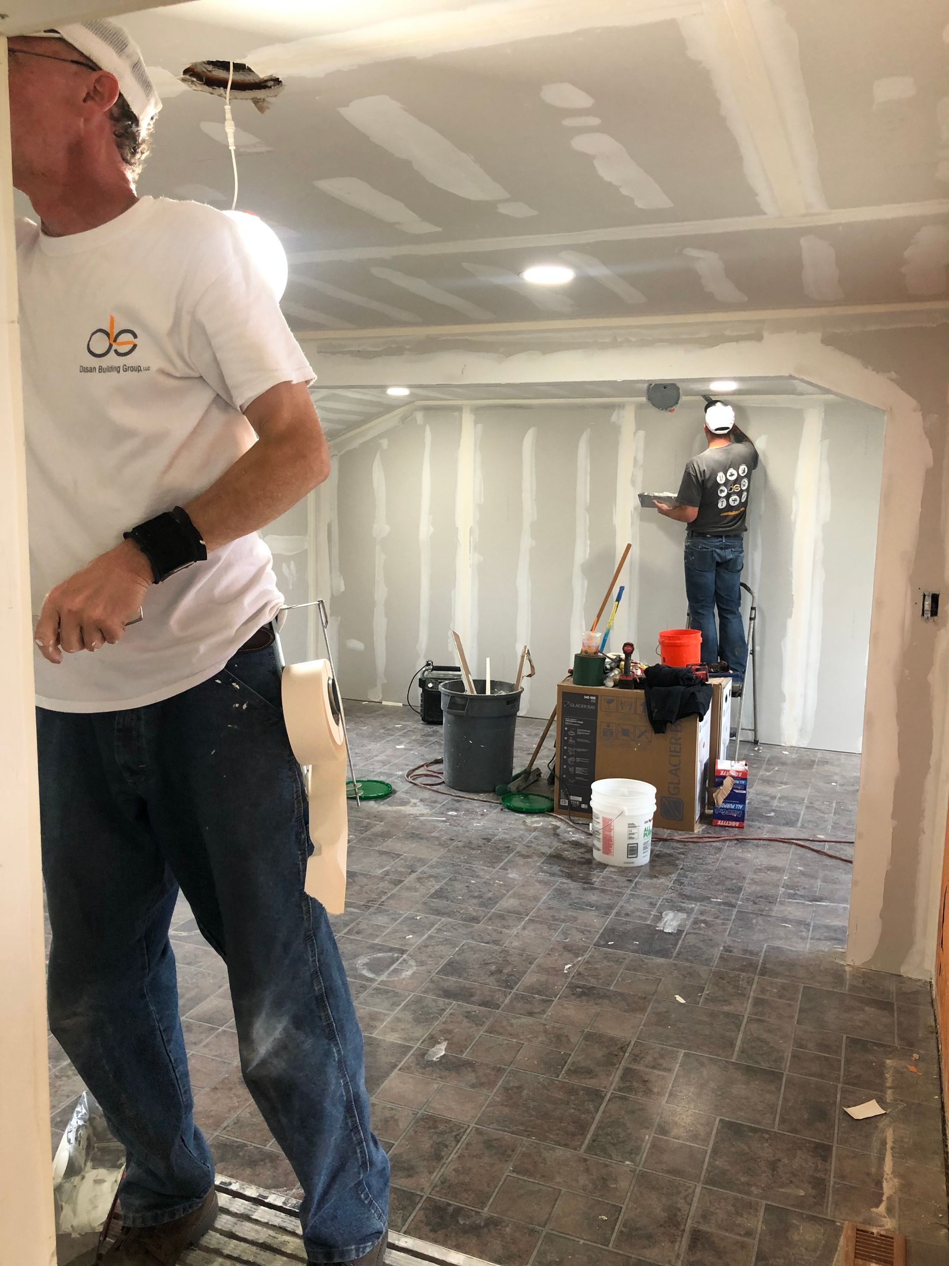 Two men drywalling a room, one on a ladder. Interior construction, tools visible.