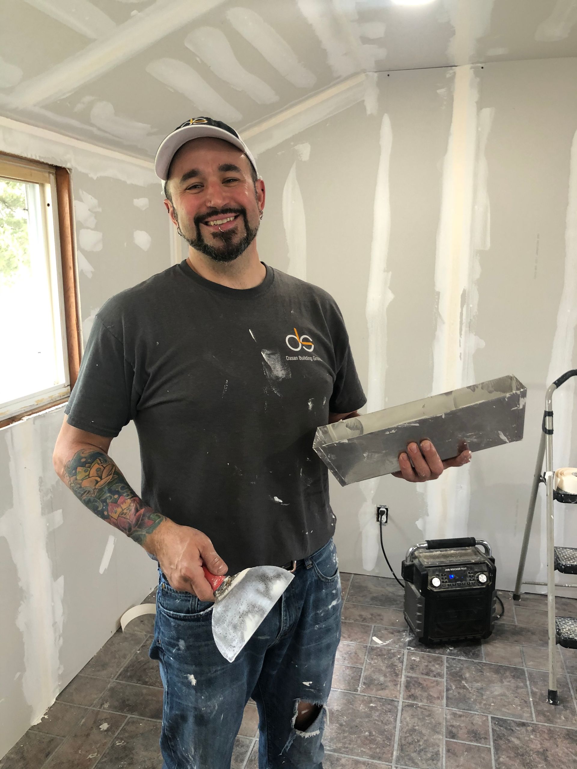 Smiling man with drywall tools in a room, wearing a cap and stained shirt.