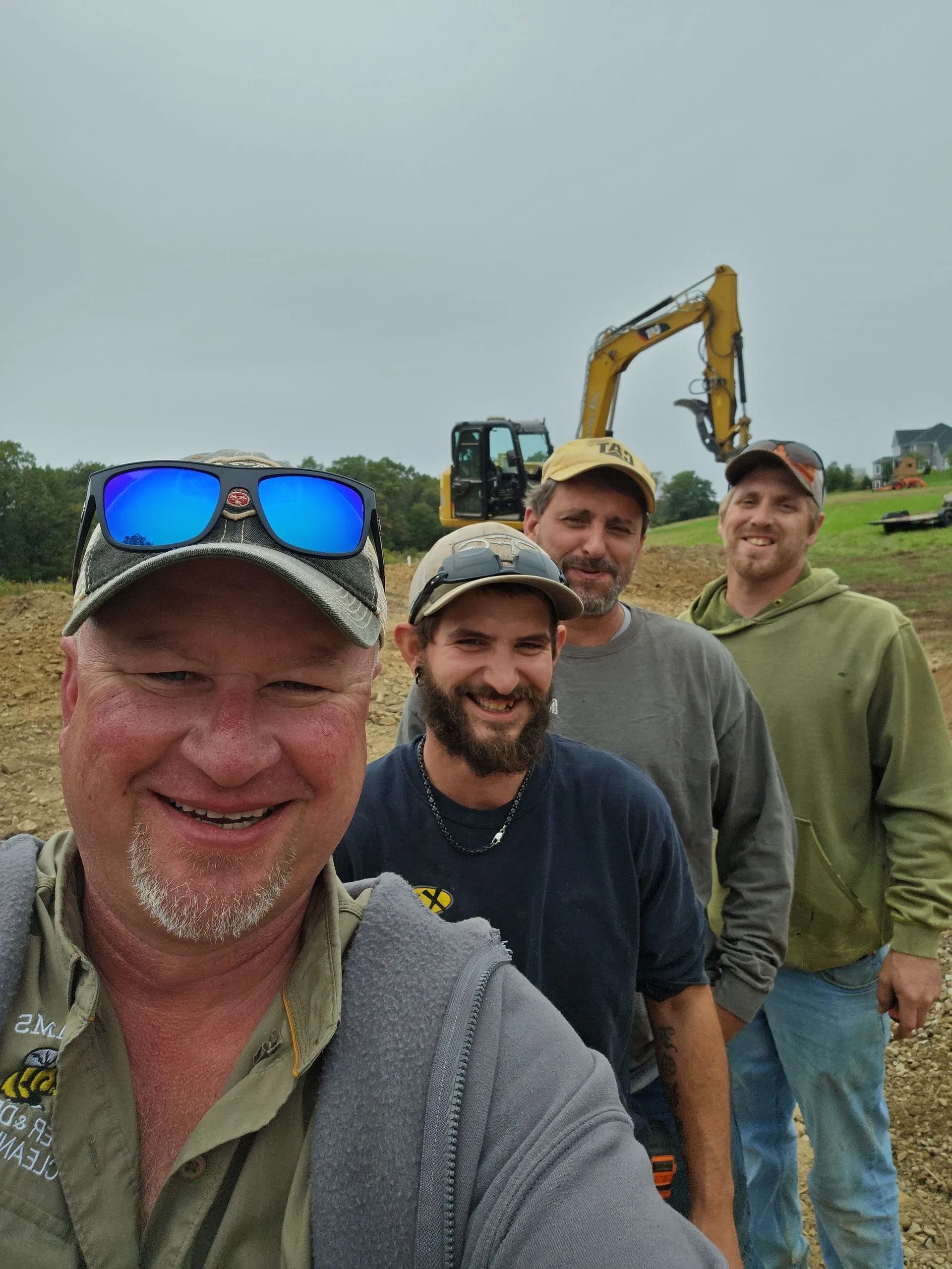 Four construction workers pose for a selfie in front of a yellow excavator on a work site.