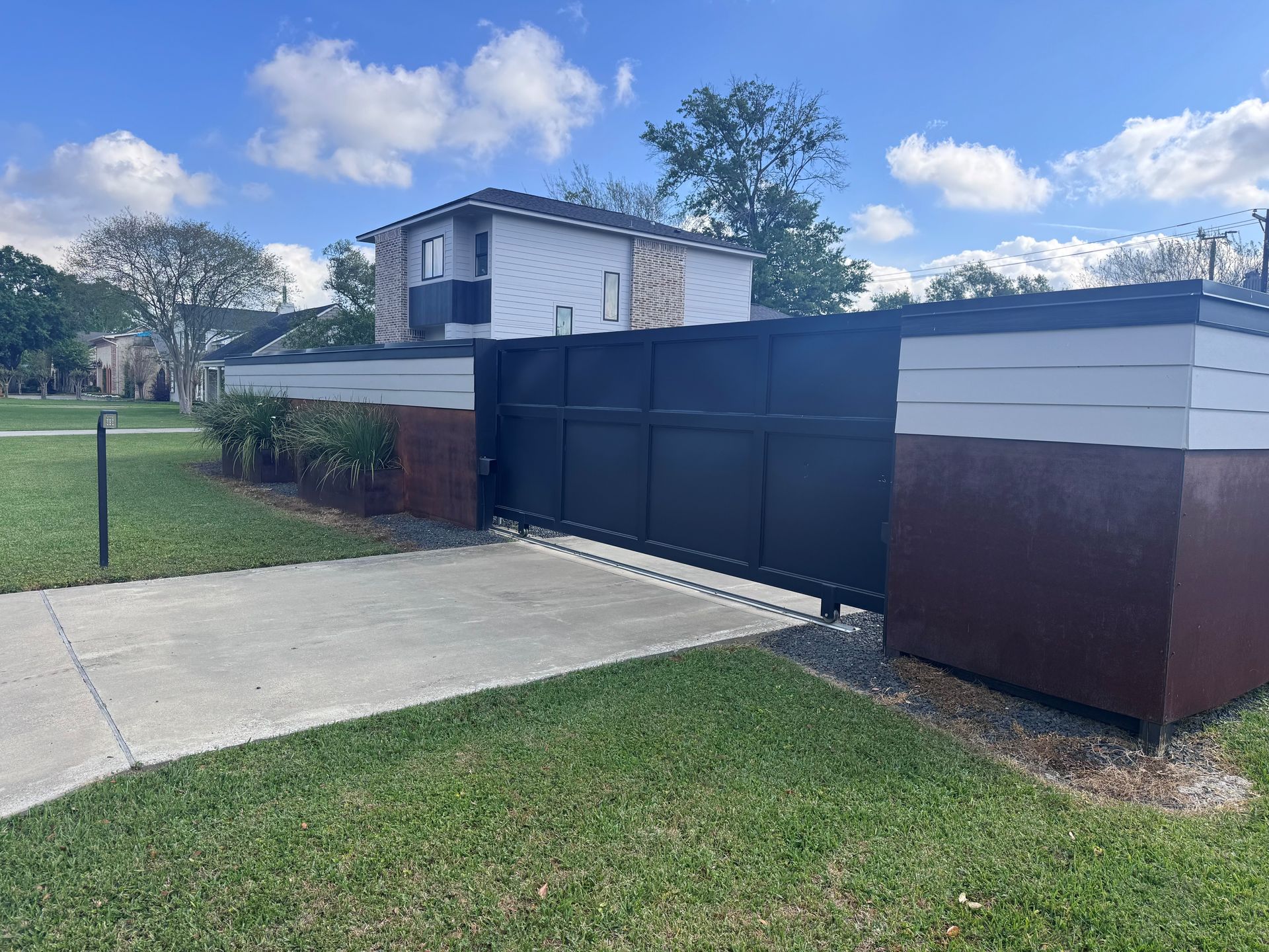 A black sliding driveway gate with dark brown and white accents, set in a grassy yard under a blue sky.