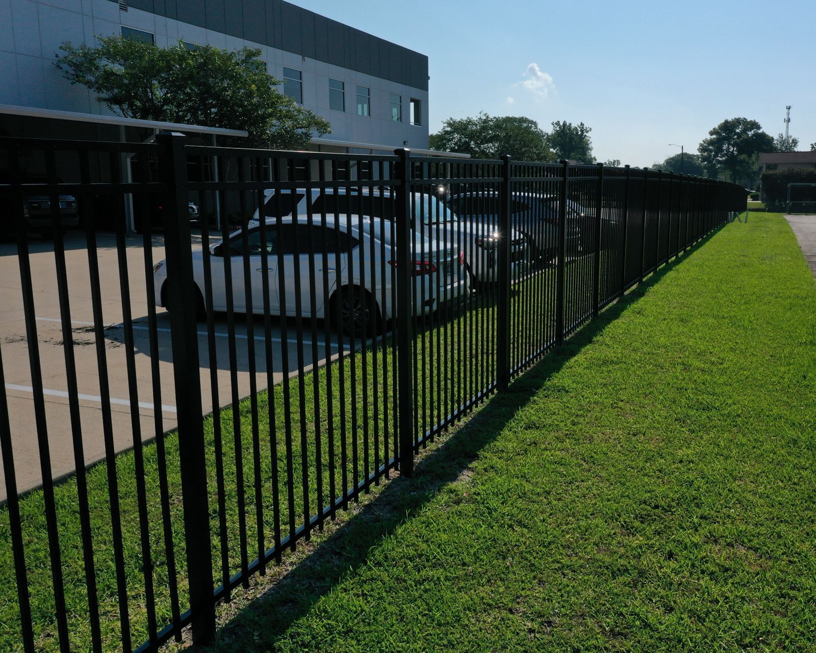 Black metal fence next to a grassy area, with cars and a building in the background on a sunny day.