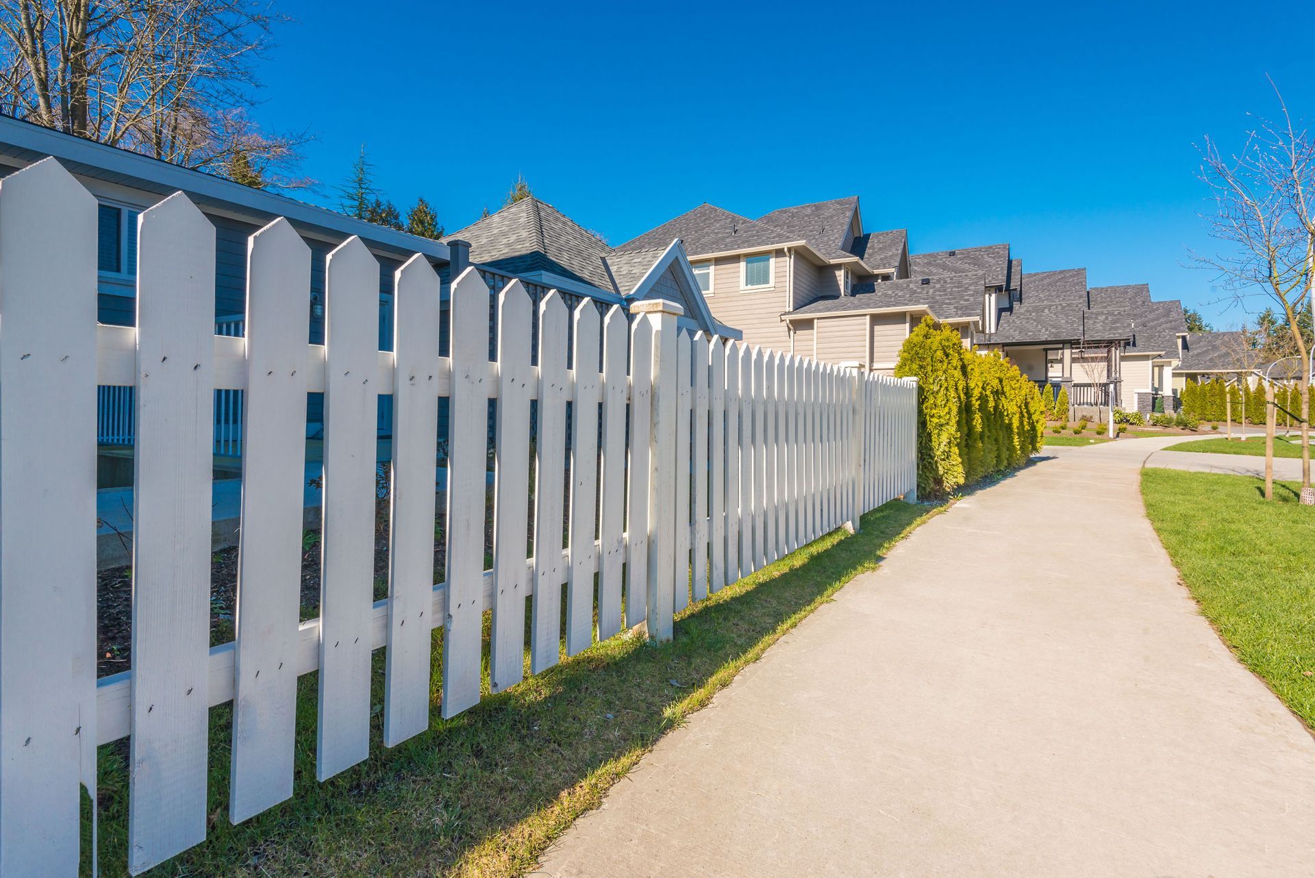 White picket fence bordering a sidewalk and houses under a clear blue sky.
