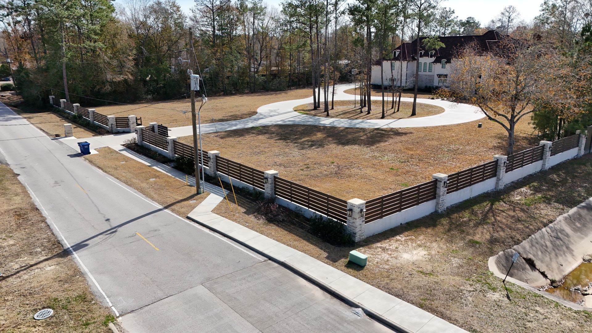 An aerial view shows a vacant lot with a circular driveway and brick fence. Trees and a house are in the background.