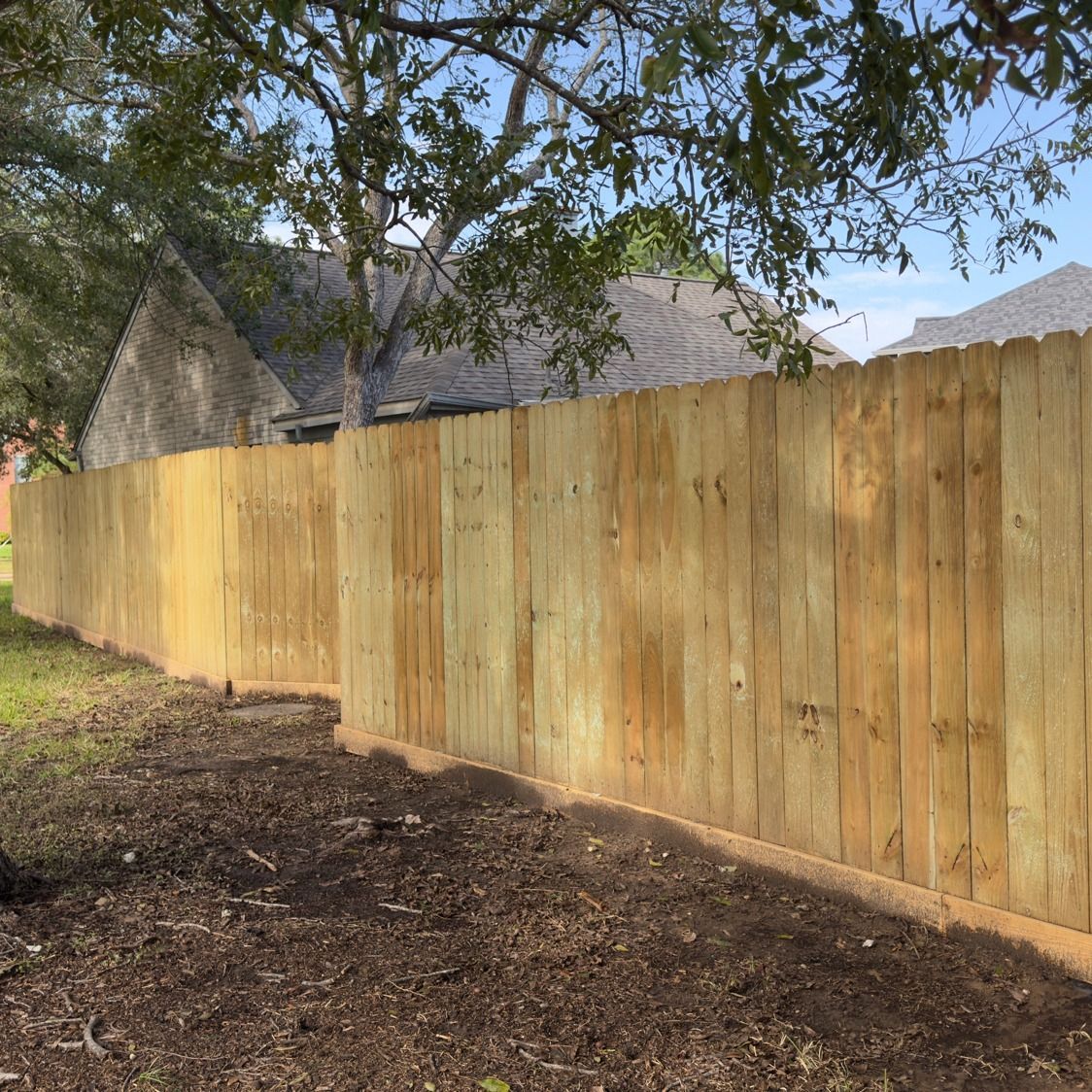 Wooden fence in a yard with brown mulch and green grass, with a house and tree in the background.