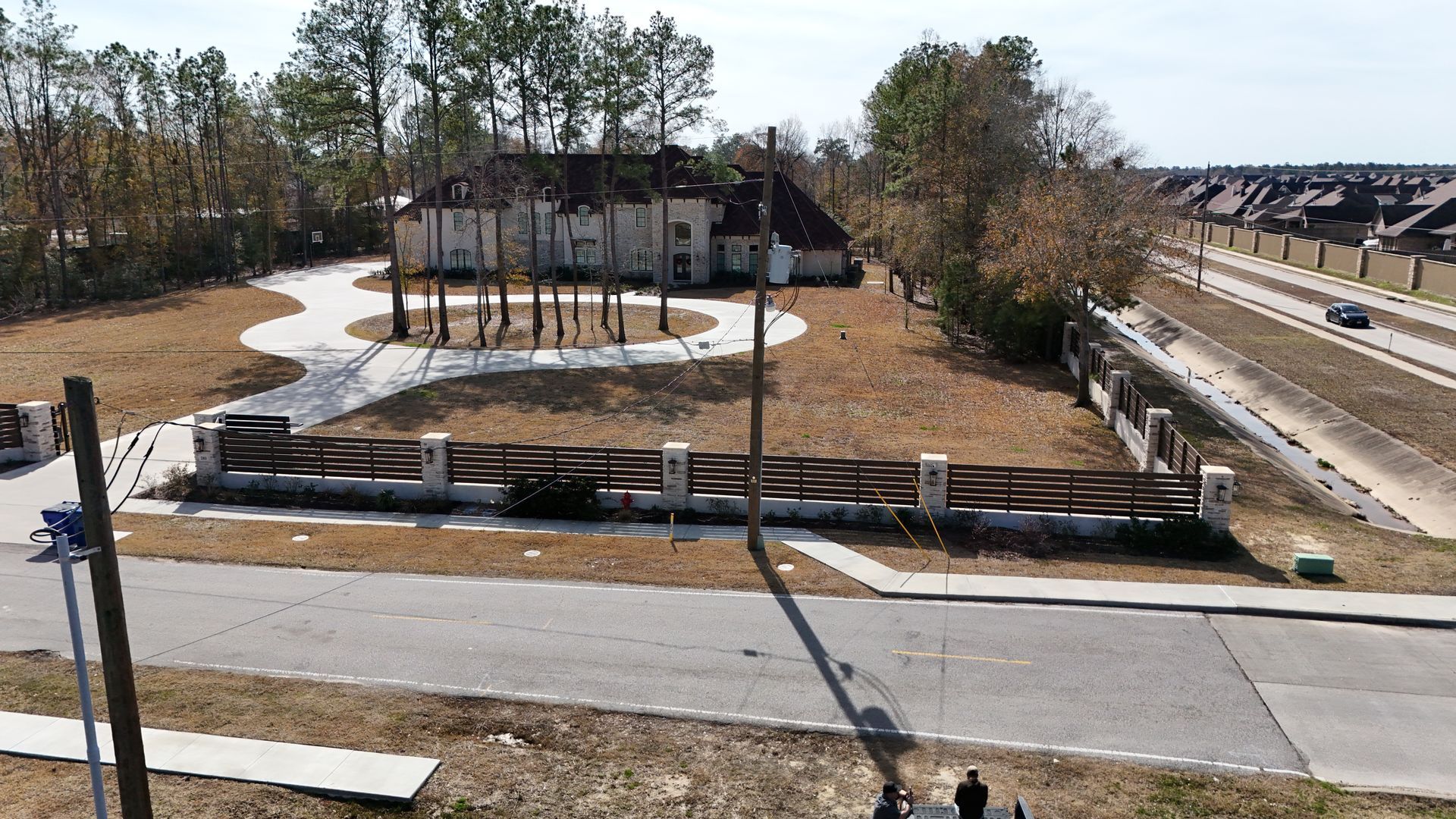 Large house with circular driveway, behind a gated fence; road and trees in view.