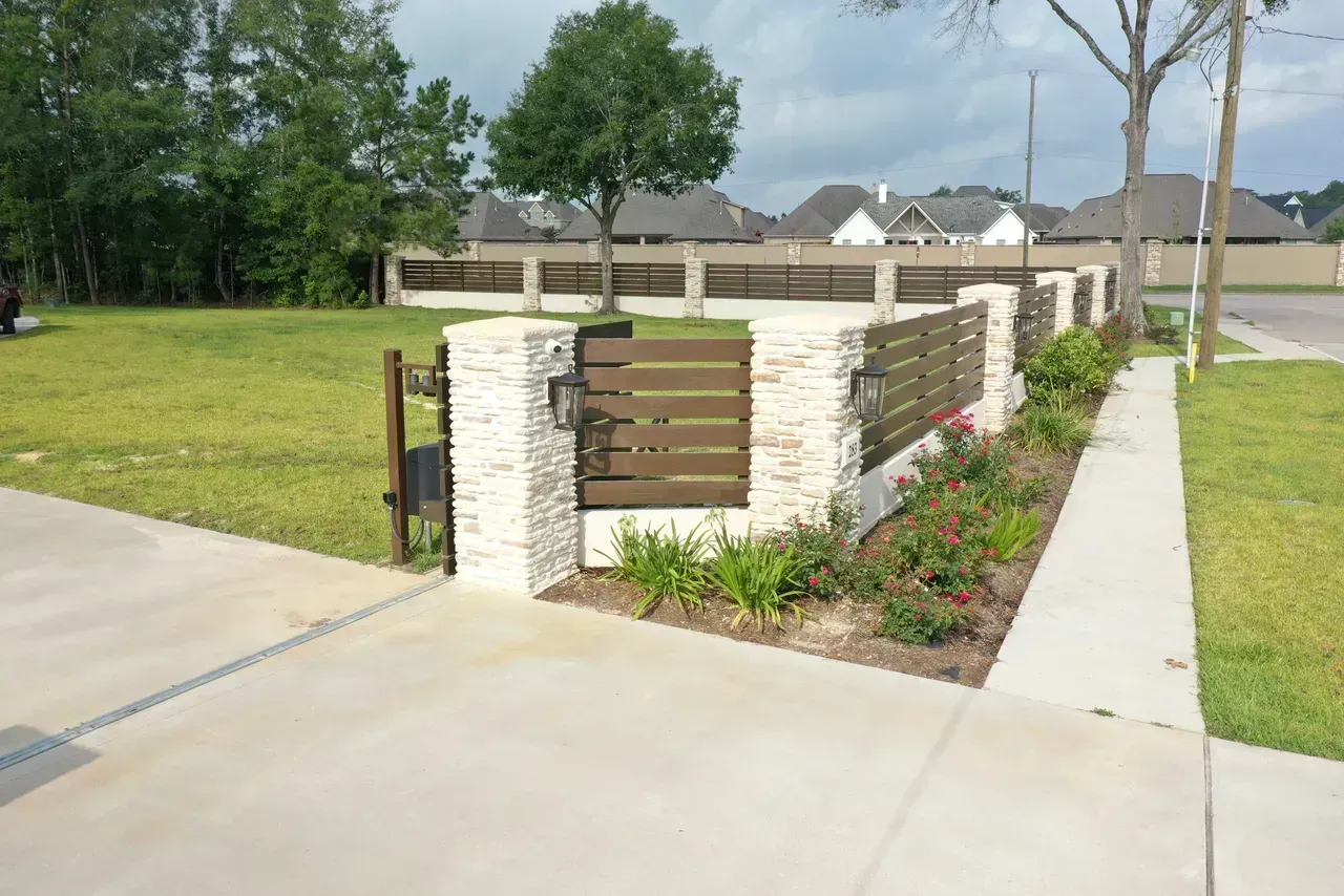 Stone and wood fence with a gate, plants, and a sidewalk in front of a green lawn and houses.
