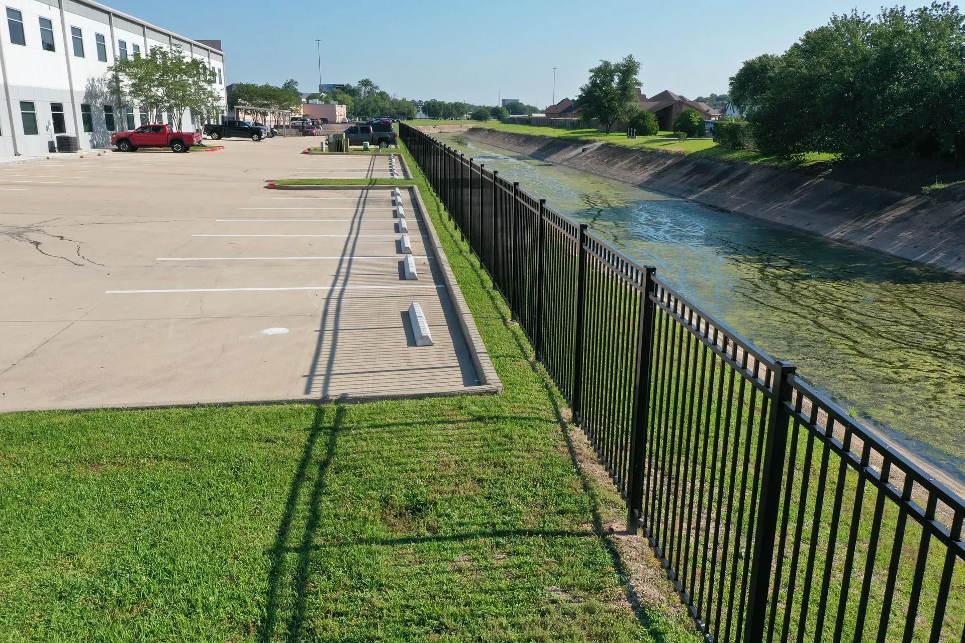 A black metal fence borders a murky waterway. Parking lot and building on the left, green grass and sky overhead.