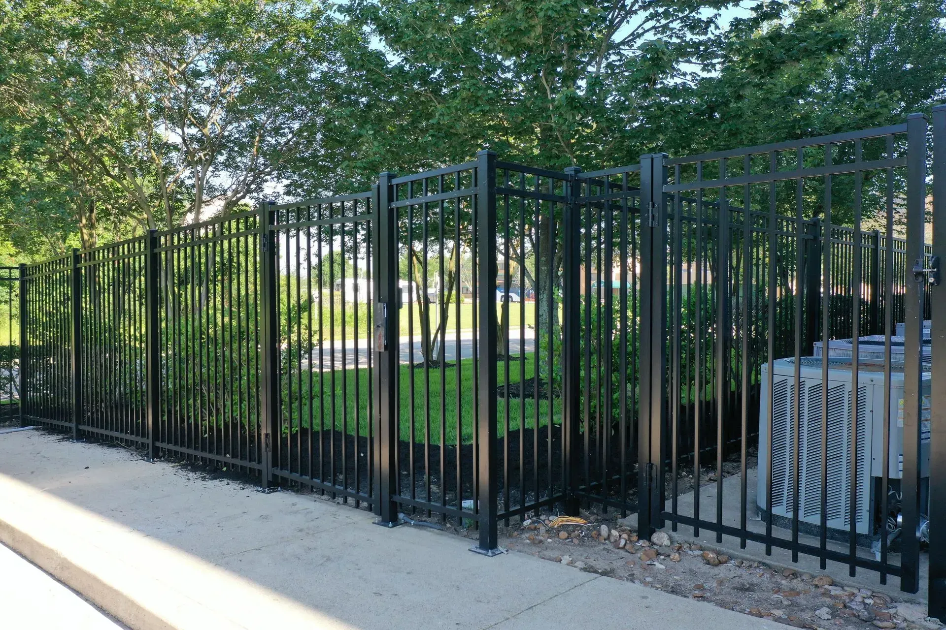 Black metal fence enclosing a small grassy area, next to an air conditioning unit and trees.