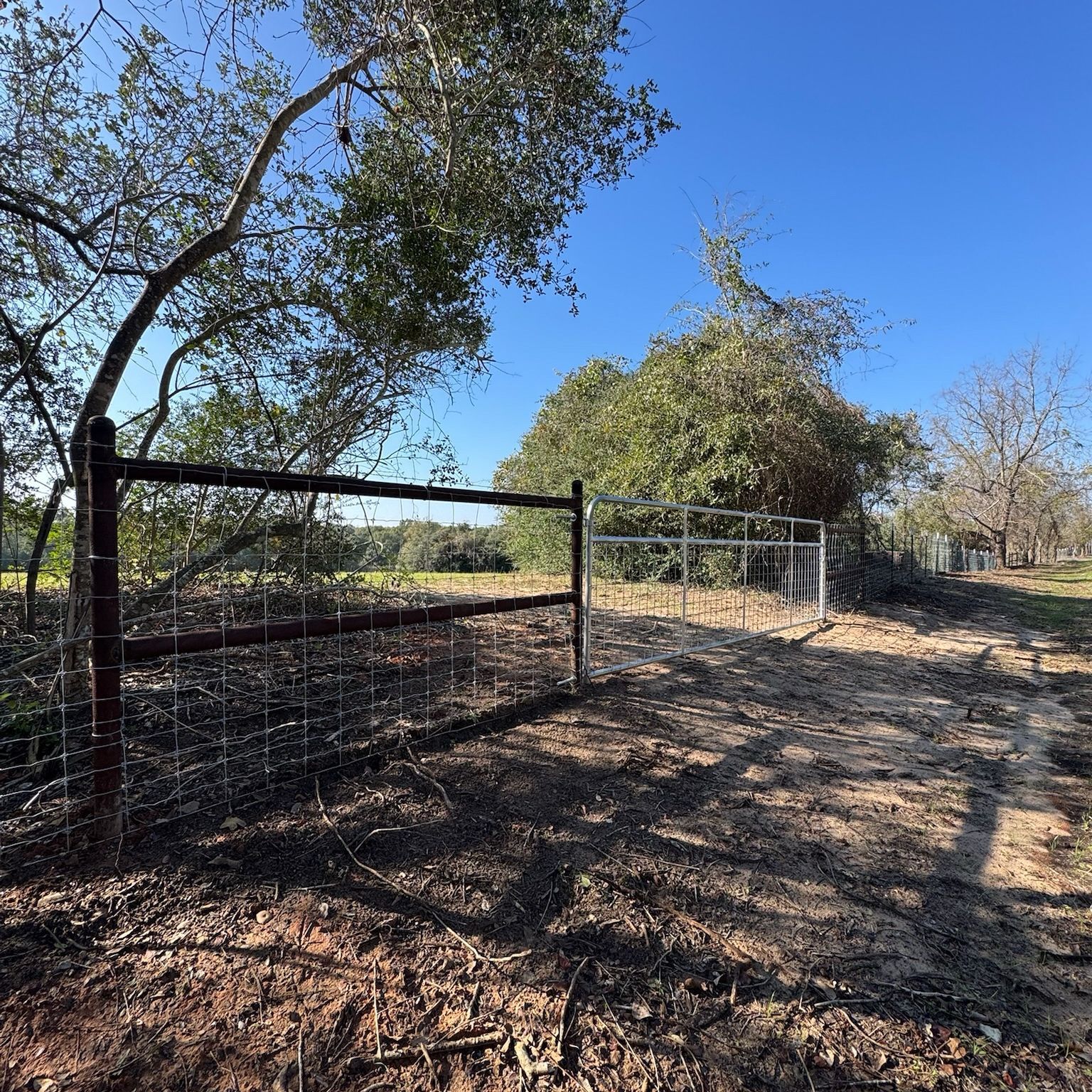 Fence gate in a rural setting, brown and silver, trees and sunny blue sky in the background.