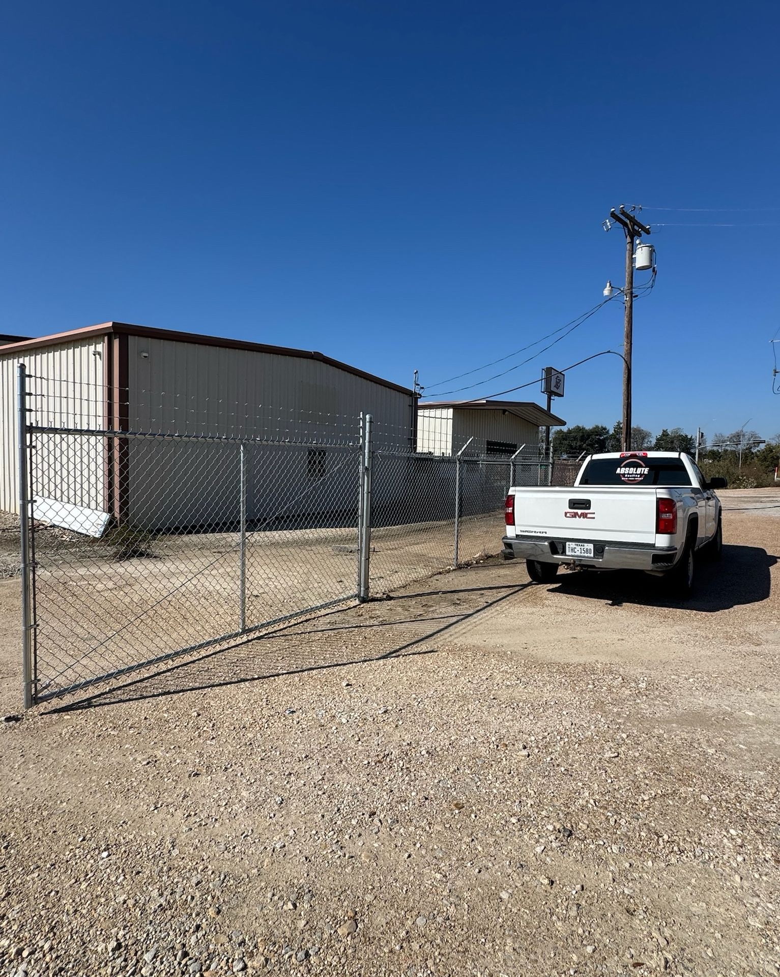 White pickup truck parked near a chain-link fence in front of a metal building on a gravel lot.