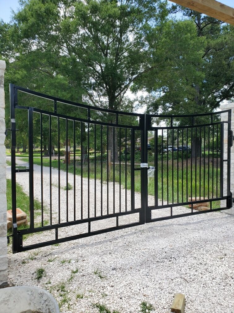 Black metal driveway gate with vertical bars, open on a gravel driveway, trees in the background.