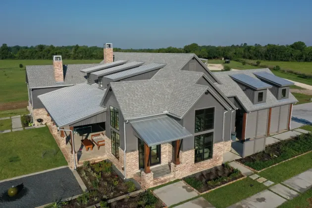 A modern gray and brick home with metal roofing sits on a grassy lot against a blue sky.