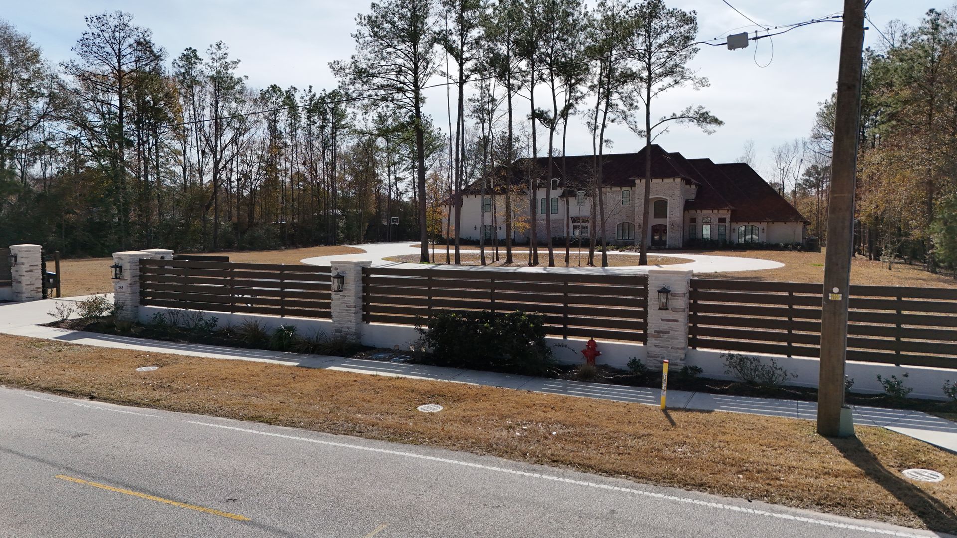 Brown horizontal slat fence with stone columns and a large house in the background.