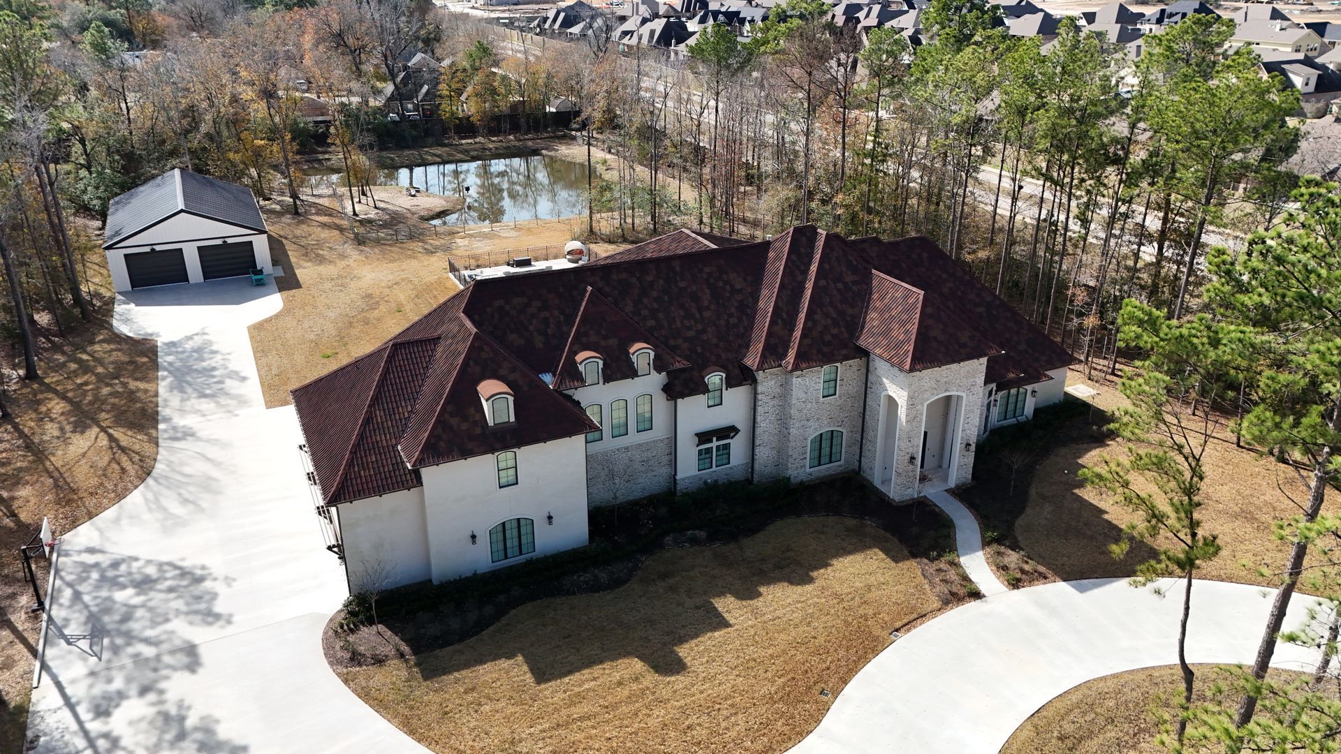 Aerial view of a large white house with a red tile roof and a detached garage on a circular driveway next to a pond.
