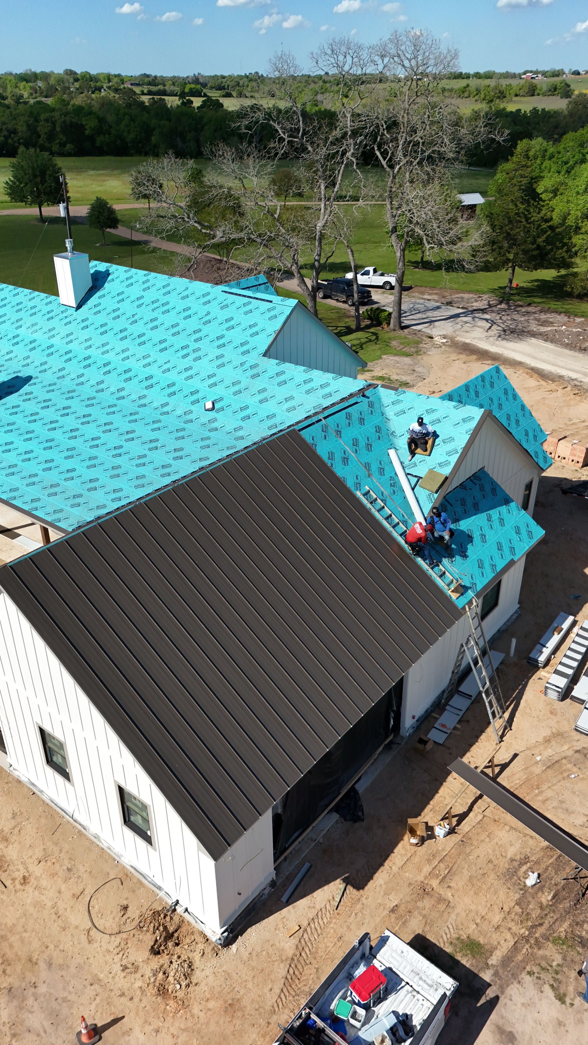 New house under construction, workers on roofs, black and blue roofing, sunny day.