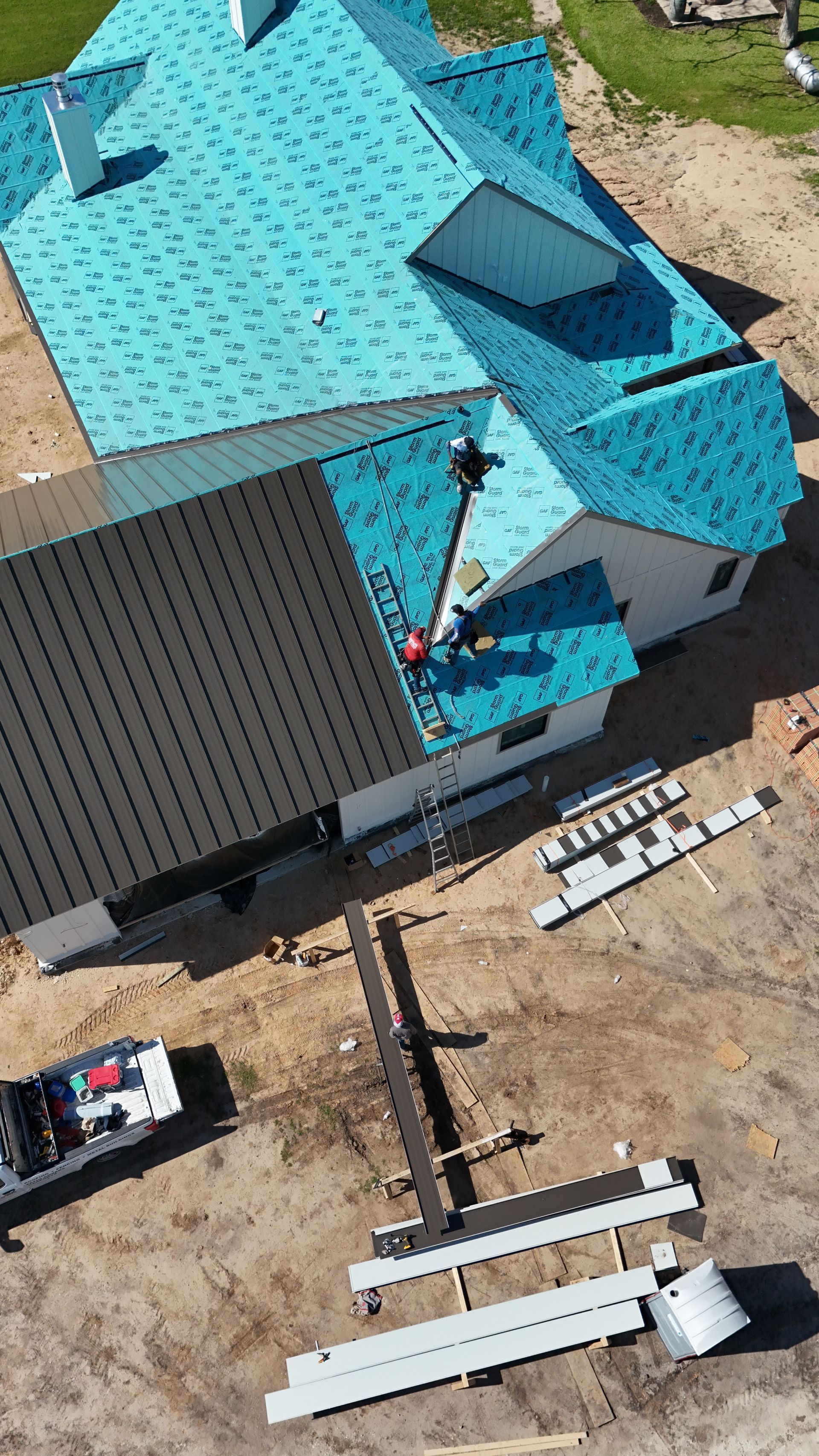 Aerial view of construction workers on a teal-roofed house with metal siding; materials and truck nearby.