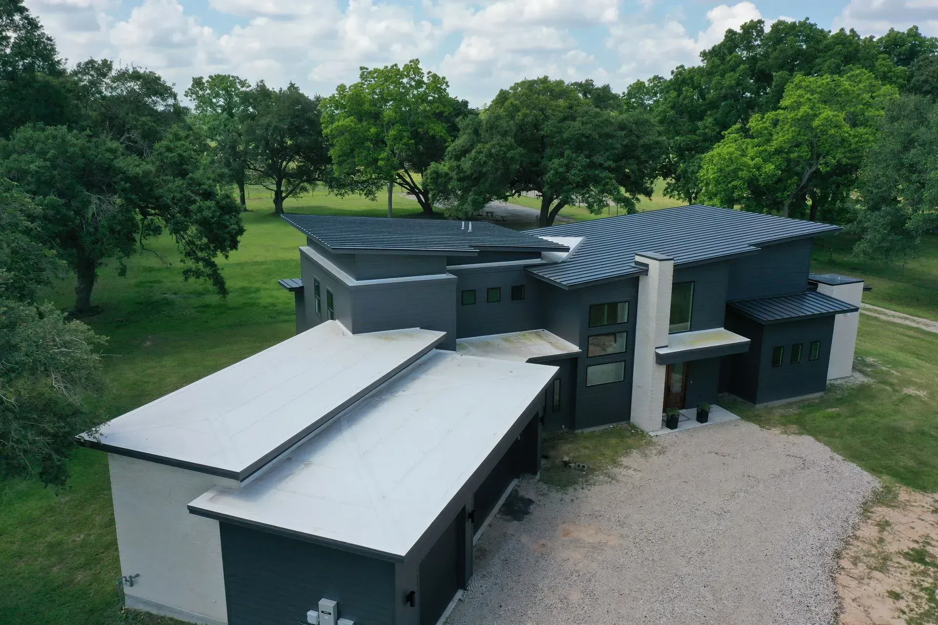 Modern gray and white house with flat silver and blue roofs surrounded by trees and green grass.