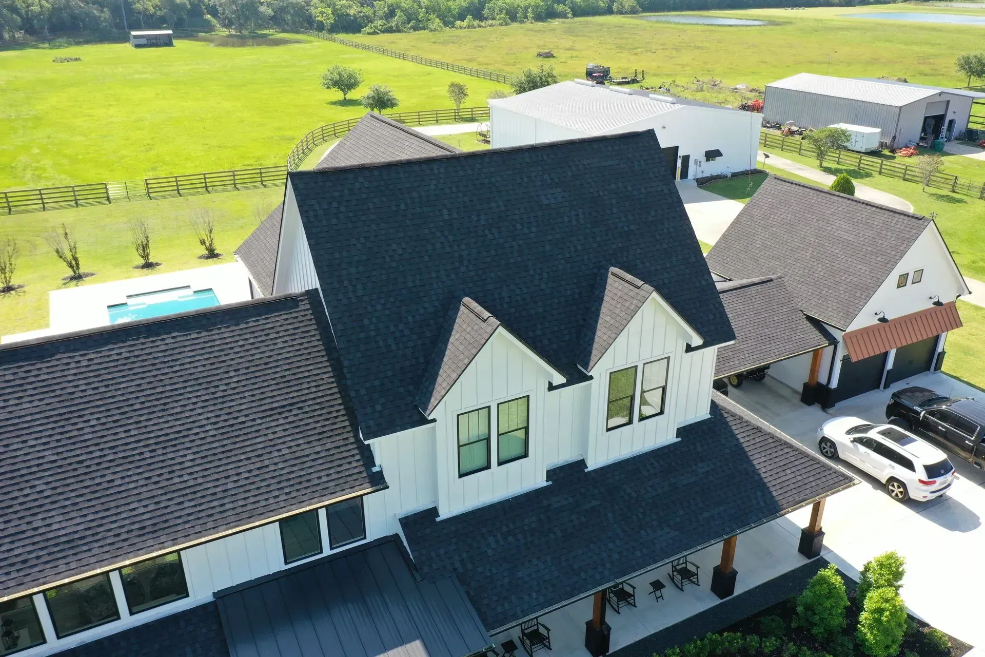 Aerial view of a white farmhouse with a black roof, a pool, and green fields in the background.