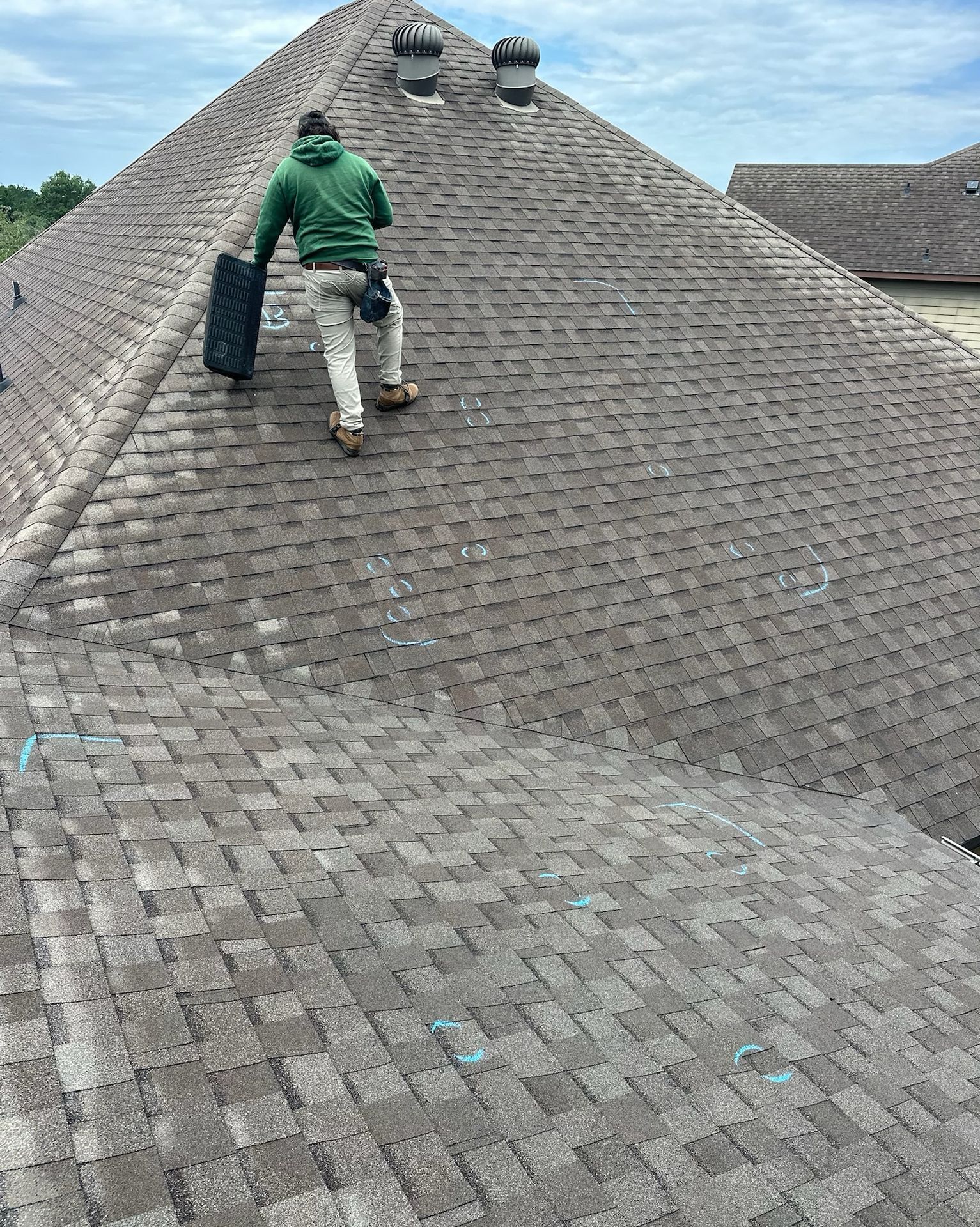 Roofer on a shingled roof, holding a black tool, cloudy sky.