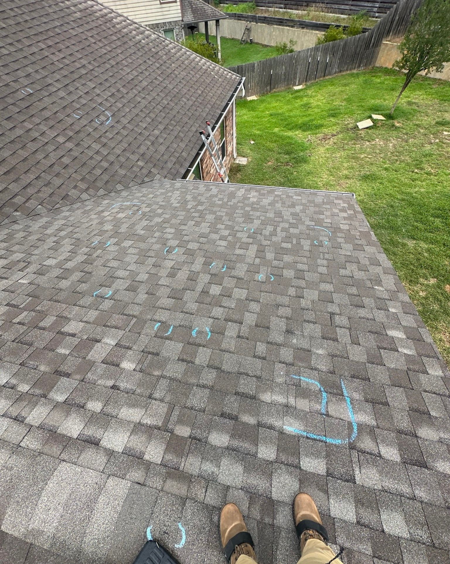 Person on a roof with blue markings on shingles, grass and fence in the background.