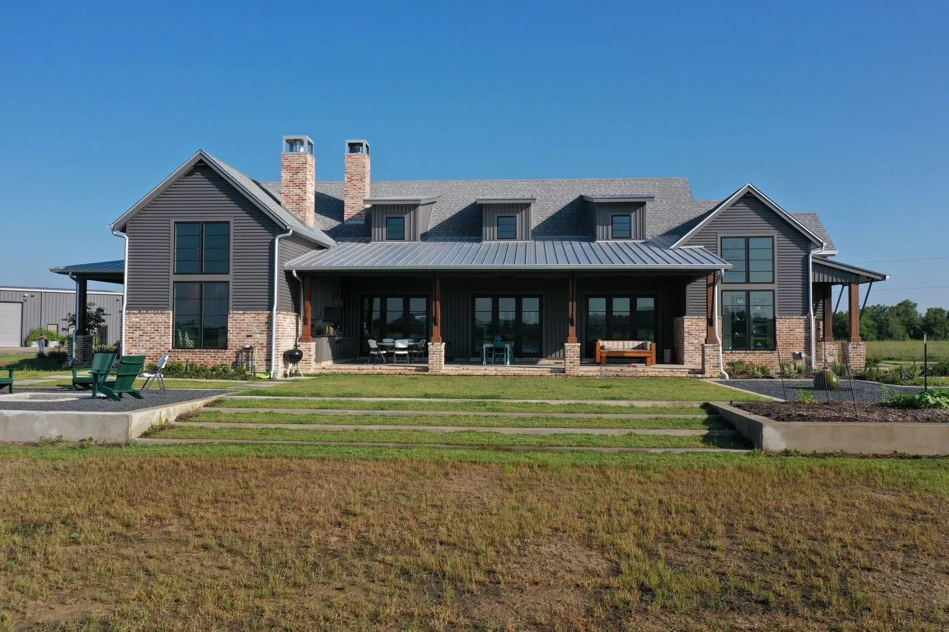 Large, two-story farmhouse with brown siding, a covered porch, and a gray roof, set against a blue sky.