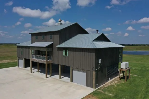 Brown, elevated house with metal roof, set on a green field under a blue sky.