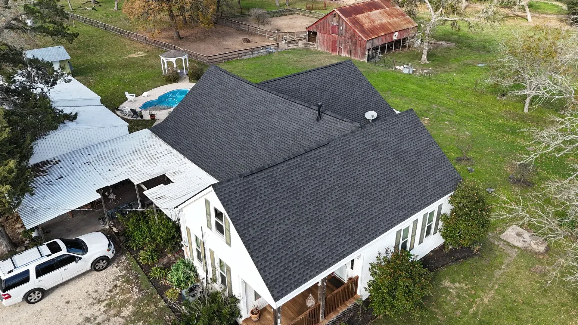White house with black roof, covered carport, white SUV, pool, and red barn in a grassy rural setting.