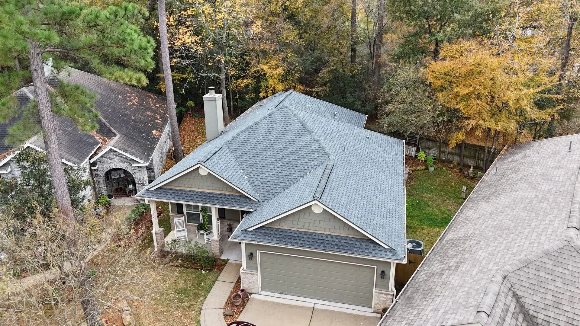 Aerial view of a gray-roofed house with a garage, surrounded by trees and a neighboring house.
