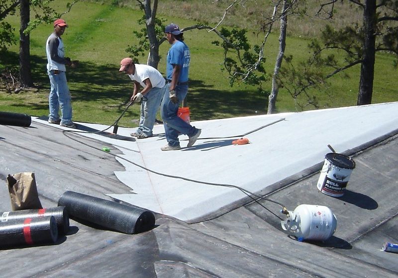 Workers applying roofing material to a sloped roof outdoors on a sunny day.